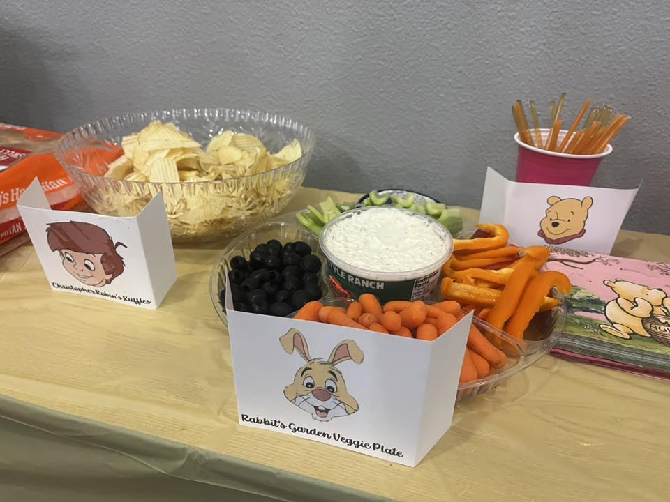 Winnie the Pooh themed snack table with veggie platter, ranch dip, and chips labeled with character signs.