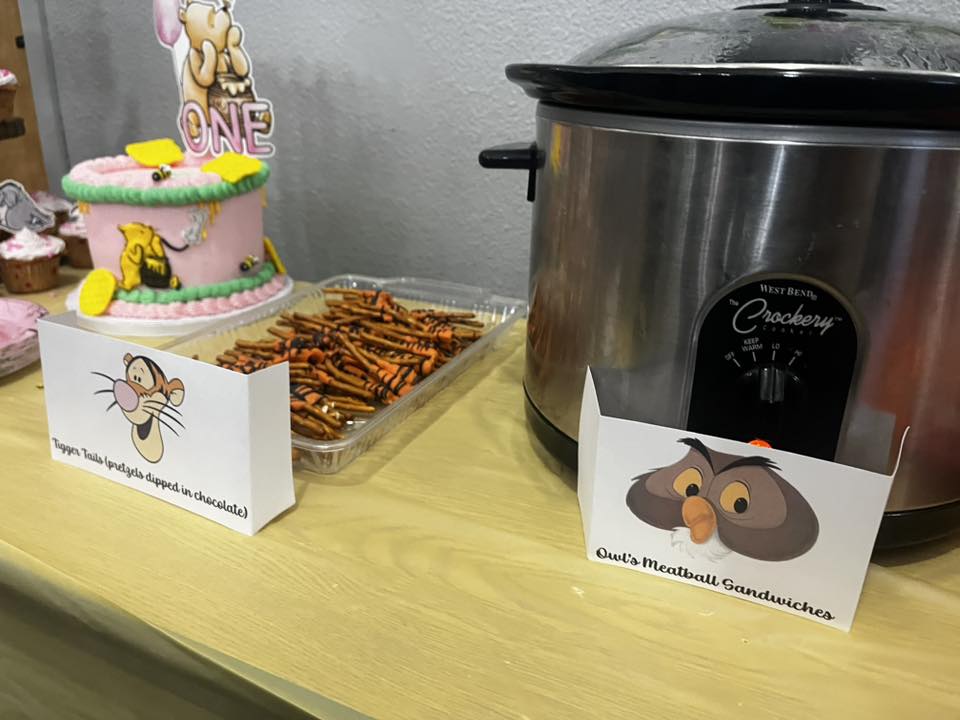 Winnie the Pooh themed party table with a cake, chocolate-dipped pretzel Tigger tails, and a slow cooker.