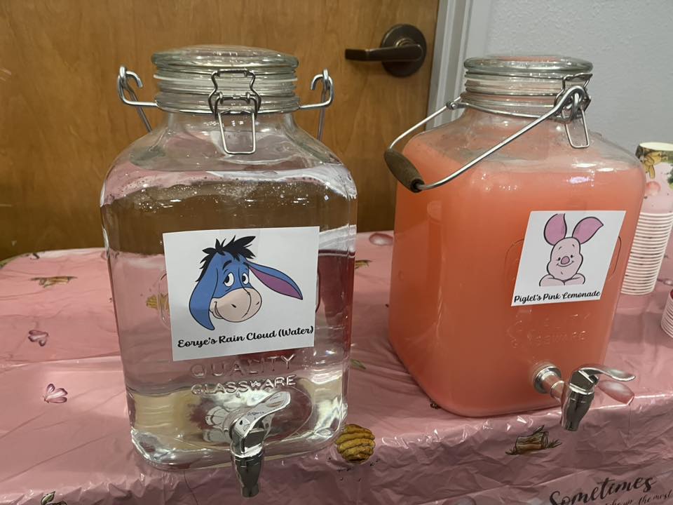 Two glass beverage dispensers labeled with Eeyore for water and Piglet for pink lemonade on a party table.