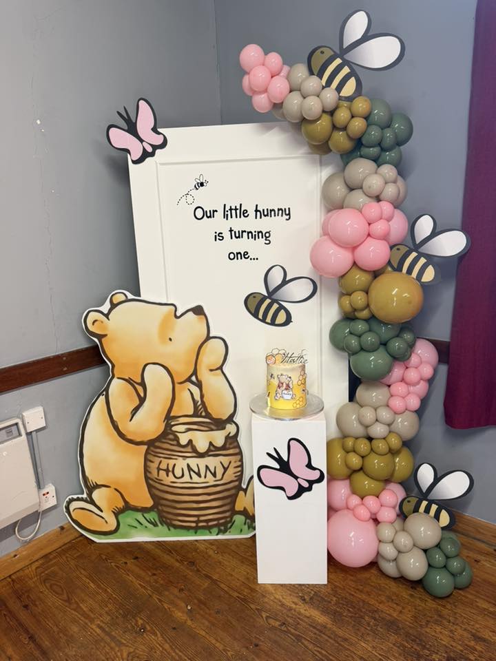 Winnie the Pooh themed first birthday party backdrop with a balloon arch, sign, and Pooh Bear cutout.