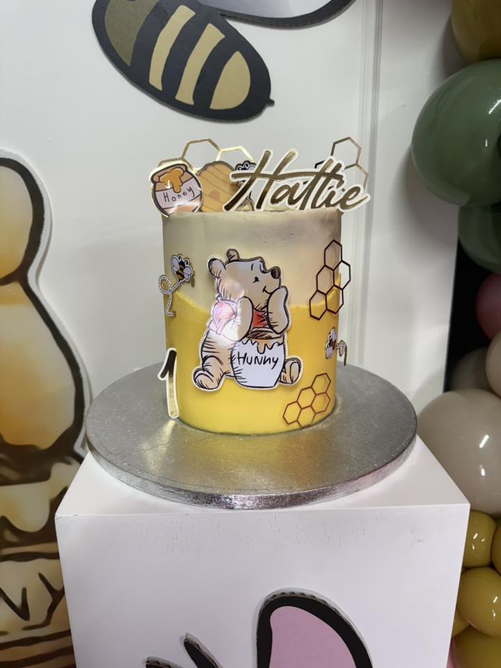 Hattie personalized Winnie the Pooh themed first birthday cake with yellow icing and honey pot decorations.