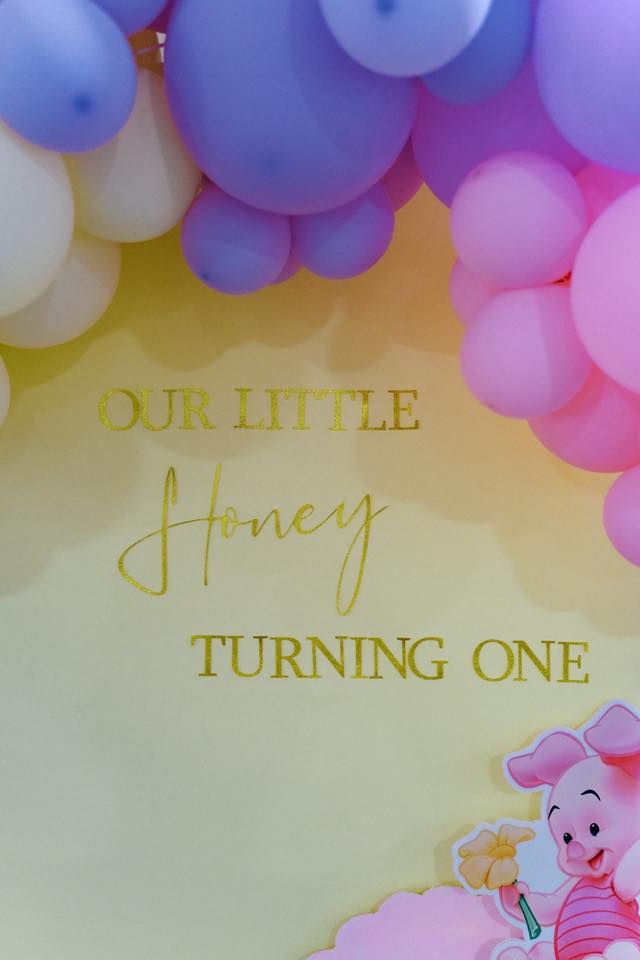 First birthday backdrop with pastel balloons and text reading Our Little Honey Turning One featuring a Piglet cutout