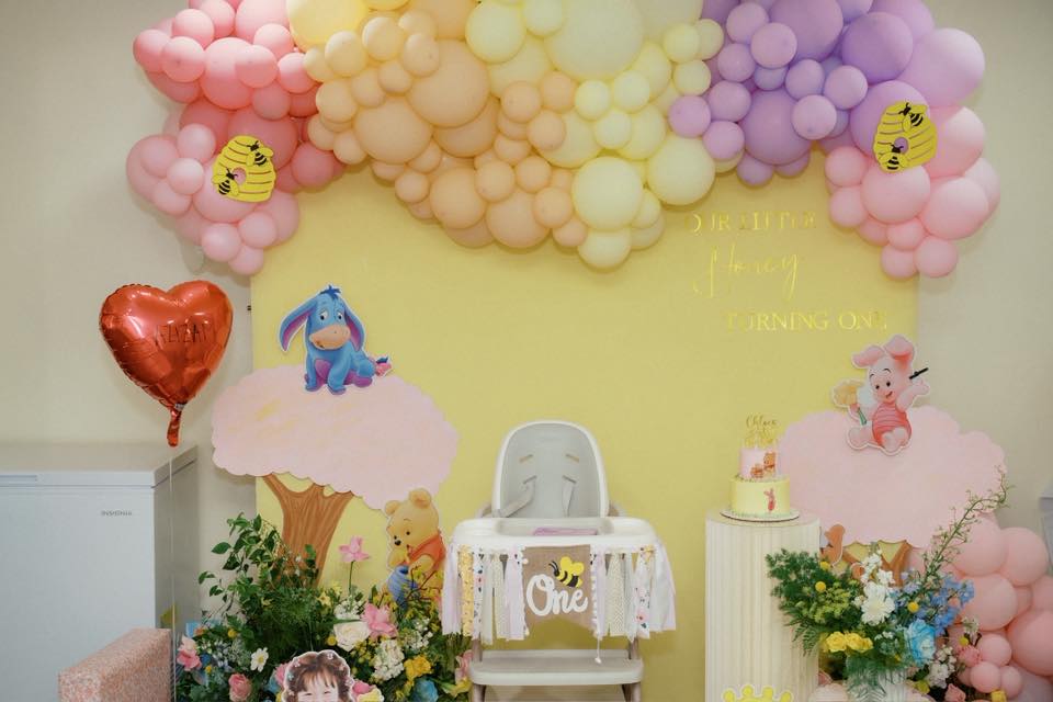 Winnie the Pooh themed first birthday party backdrop with a balloon arch, high chair, and decorative signage.