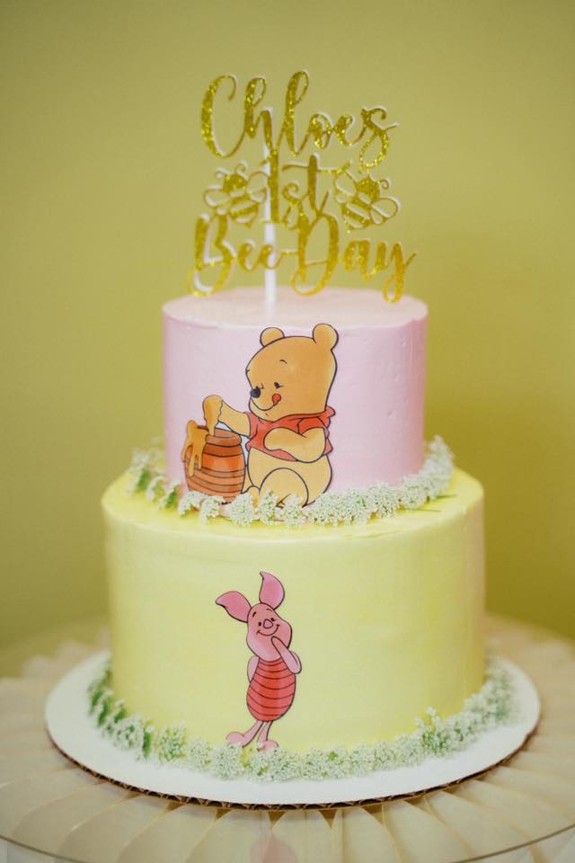 Two-tiered pink and yellow Winnie the Pooh themed first birthday cake with a Chloe's 1st Bee Day glitter topper