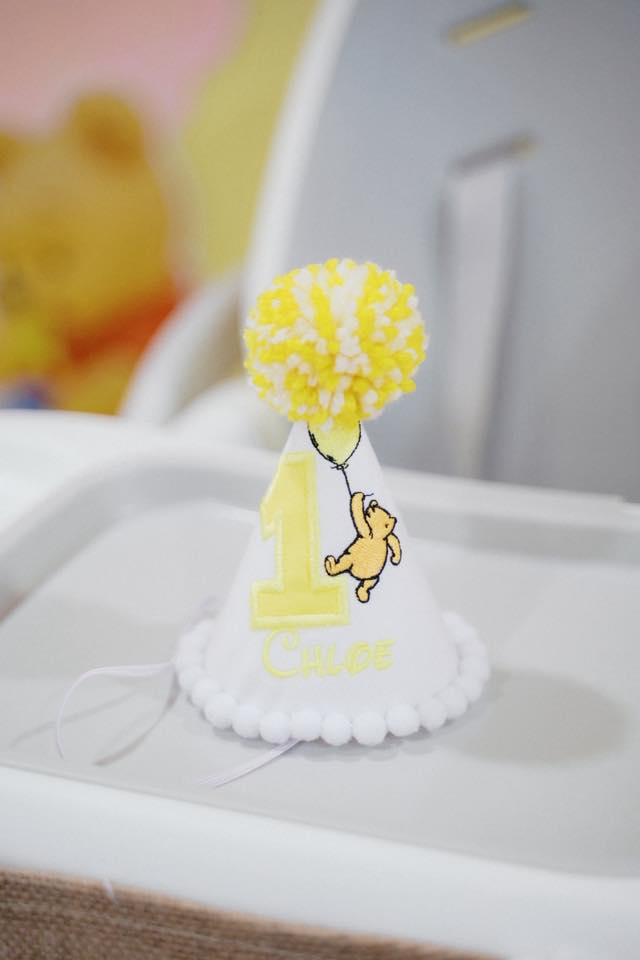 Personalized Winnie the Pooh inspired first birthday party hat with yellow pom-pom and embroidered name.