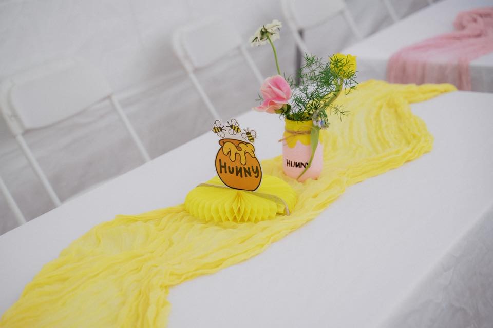 Table setting with yellow honey-themed decorations, including a Hunny pot sign, flower jar, and yellow table runner.