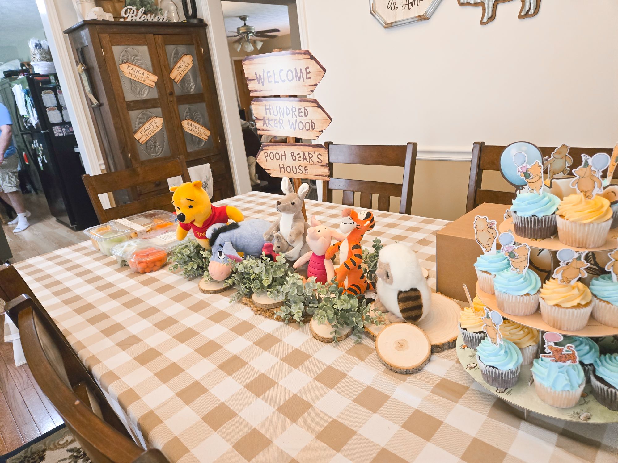 Winnie the Pooh themed party table centerpiece featuring character plushies, a wooden Hundred Acre Wood sign, cupcake tower.