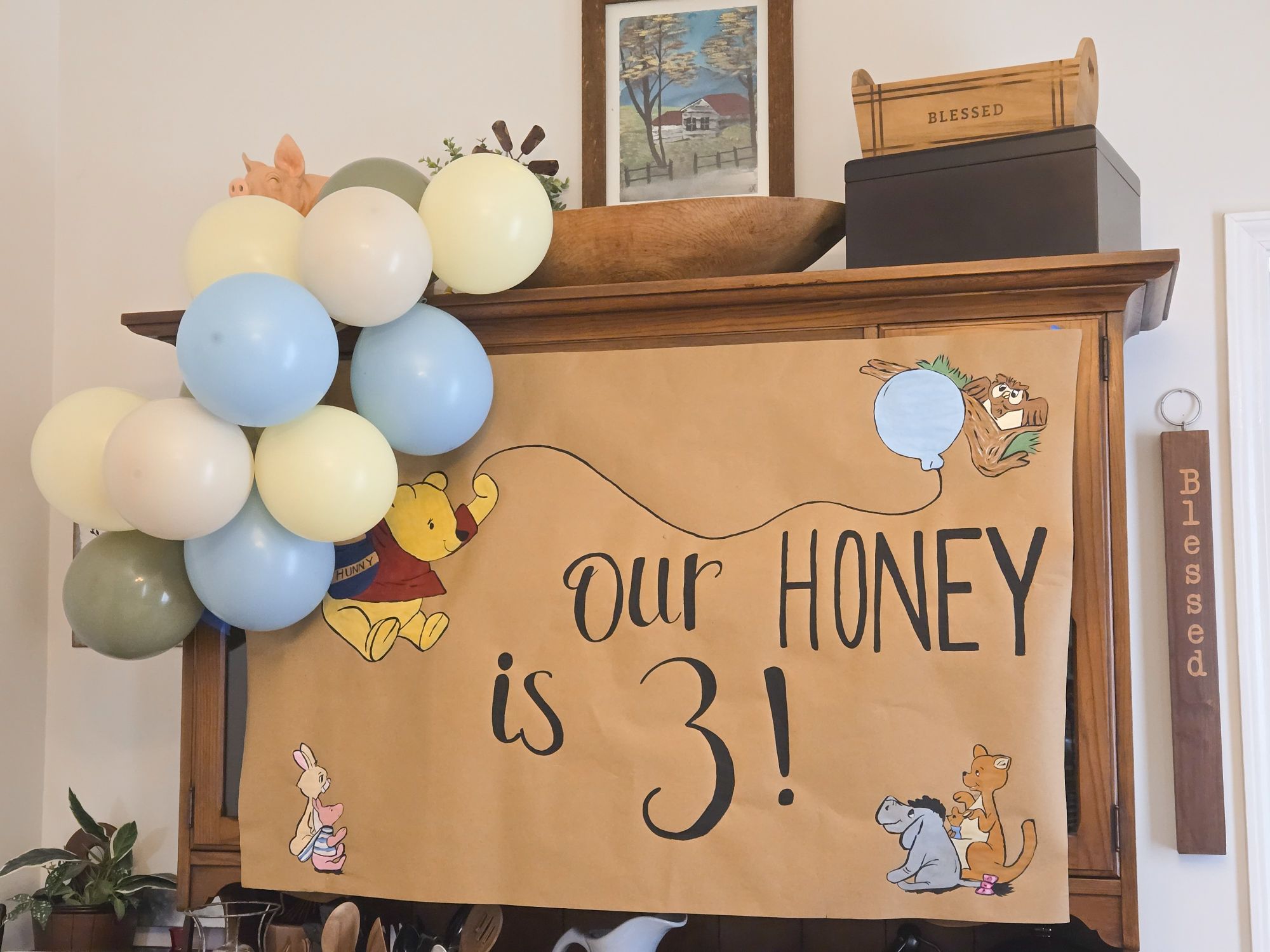 Handcrafted Winnie the Pooh themed birthday sign reading Our Honey is 3, decorated with a balloon garland.