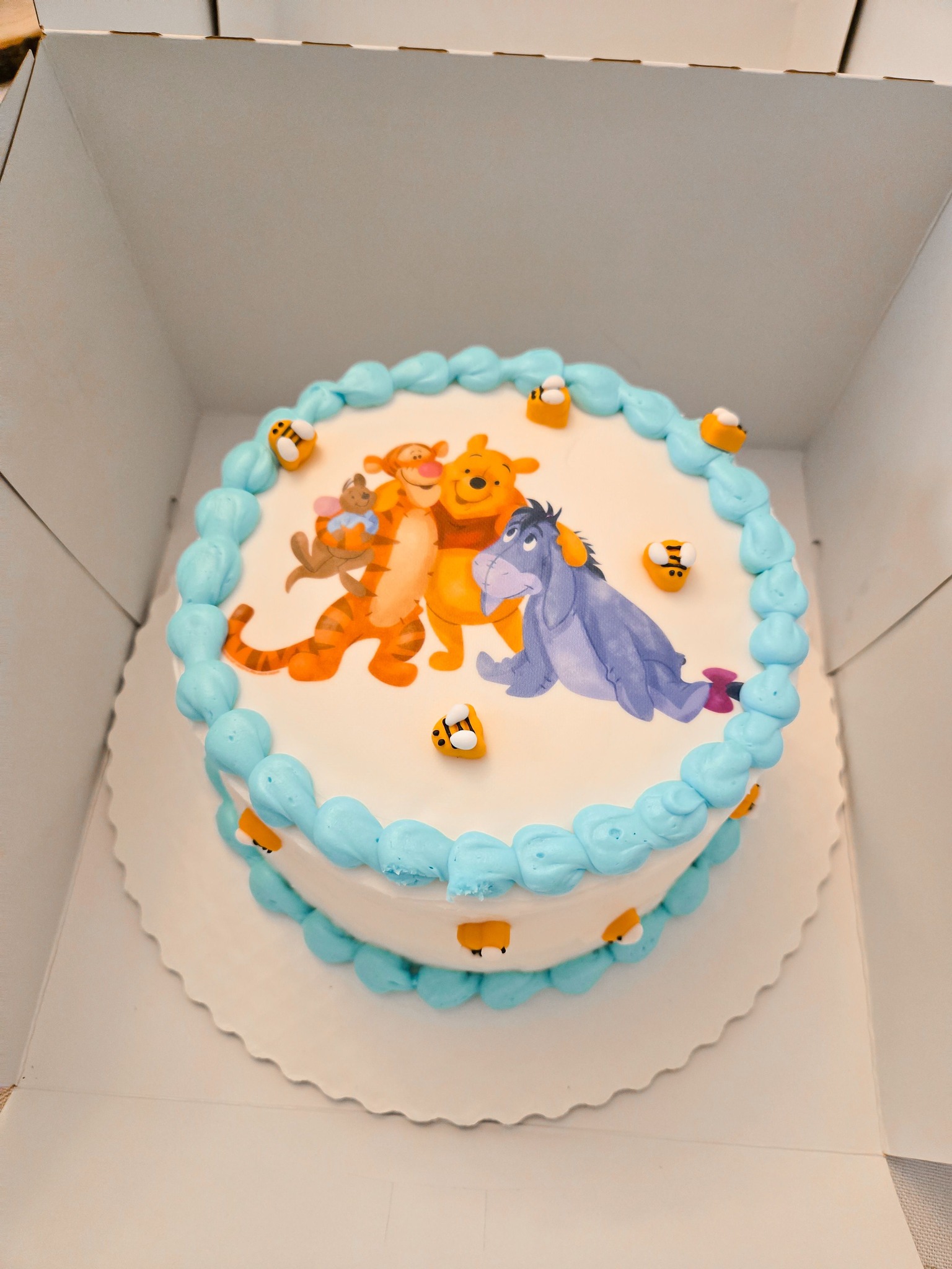 Round cake featuring Winnie the Pooh, Tigger, and Eeyore with light blue frosting and bee decorations.