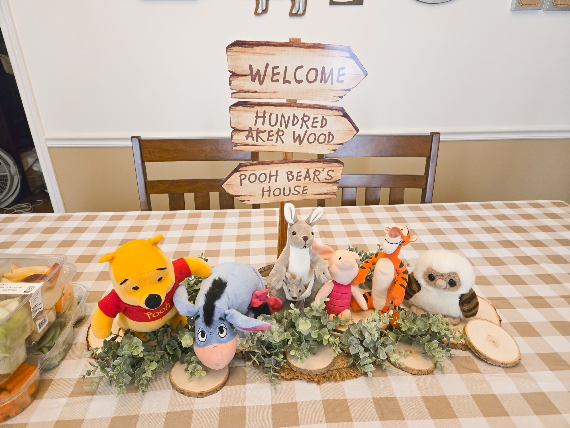 Winnie the Pooh and friends plush toys gathered around a Hundred Aker Wood welcome sign on a dining table.