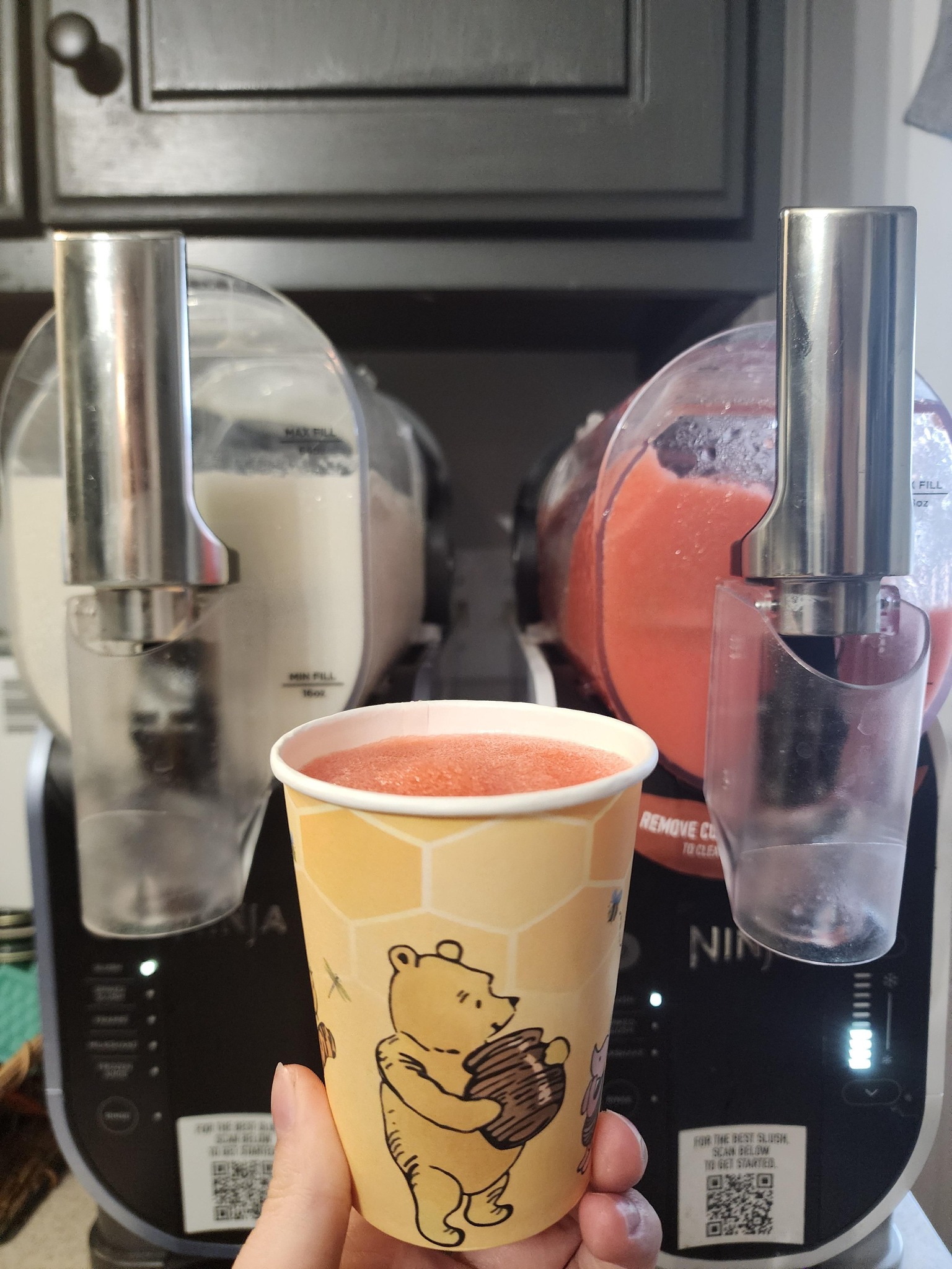 Hand holding a Winnie the Pooh paper cup filled with a frozen red strawberry slushie in front of a dual-chamber Ninja slushie machine.
