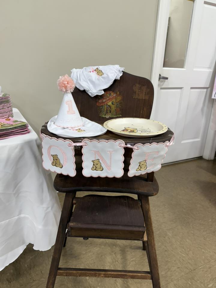 Wooden high chair decorated with Winnie the Pooh 'ONE' banner, party hat, and matching plate for a first birthday.