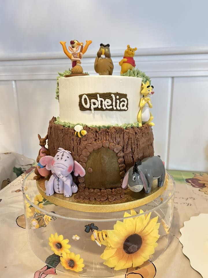 Two-tier Winnie the Pooh themed birthday cake featuring the characters and a tree trunk base personalized with the name Ophelia.