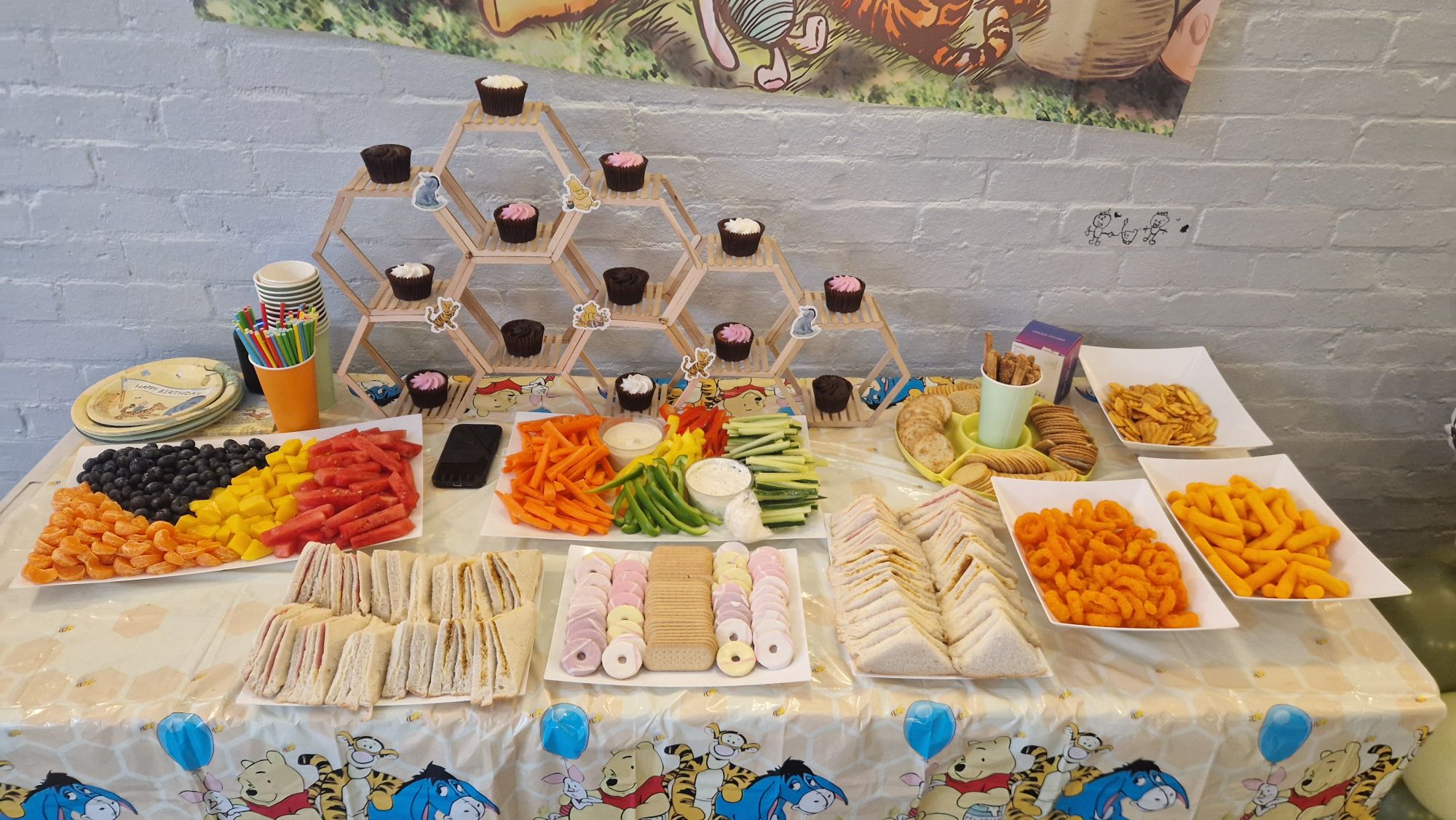 Winnie the Pooh themed party table with cupcakes, finger sandwiches, fresh fruit, crackers, and snacks.