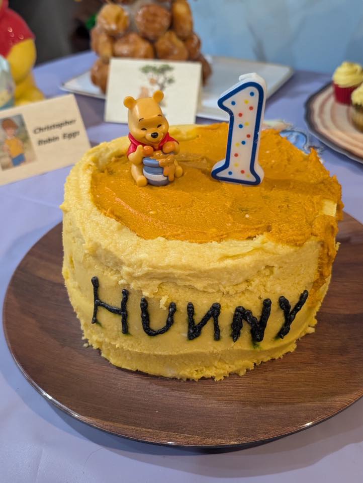 Winnie the Pooh first birthday cake with yellow icing, a Hunny pot topper, and a number one candle.