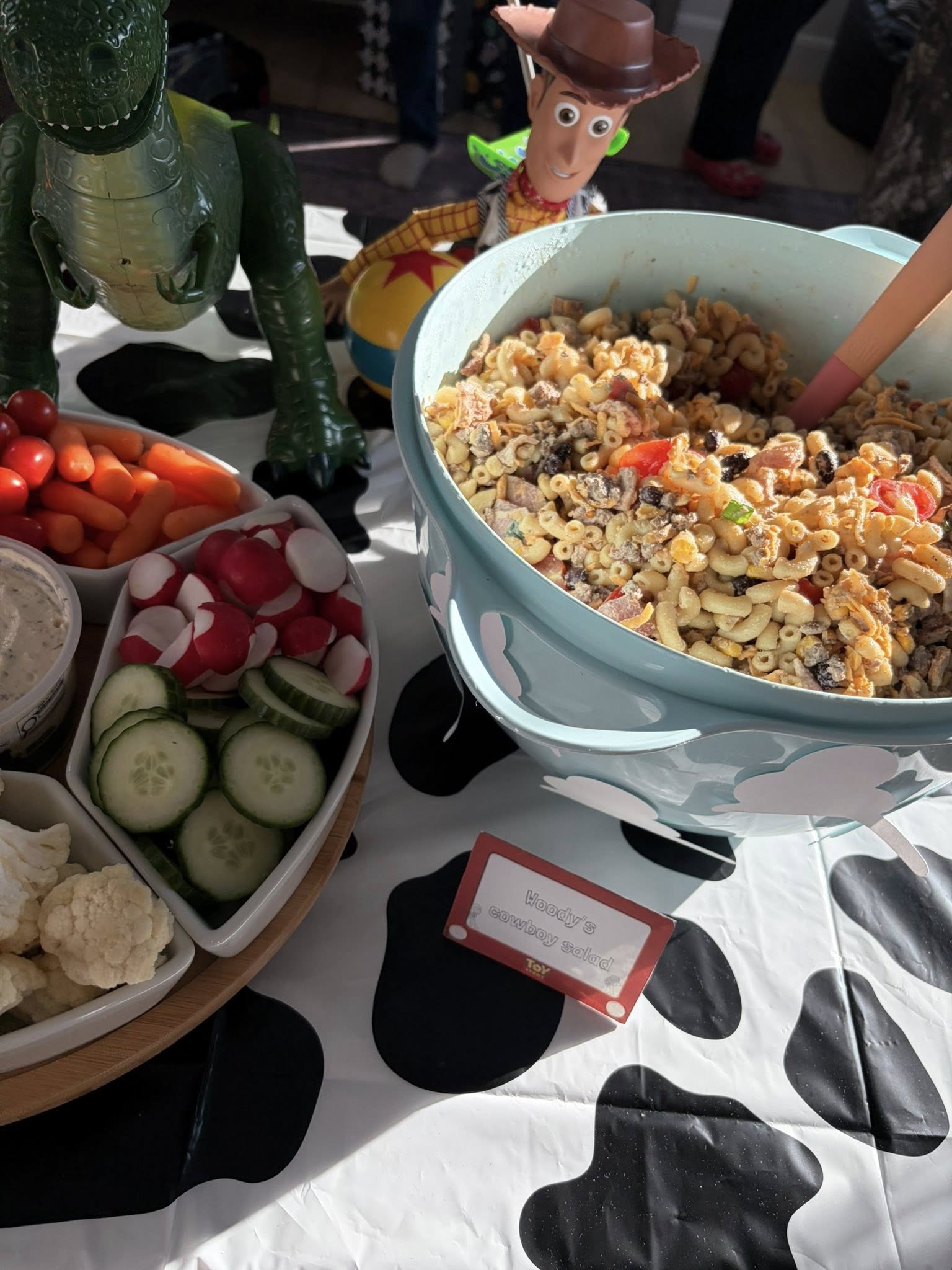 Bowl of pasta salad labeled Woody's cowboy salad surrounded by vegetable snacks and Toy Story themed party decor.