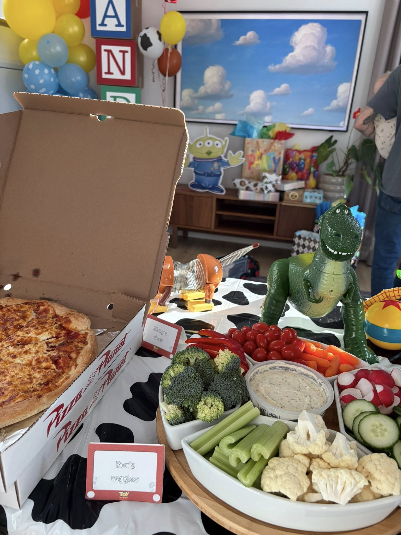 Toy Story themed party table with Pizza Planet pizza box, assorted fresh raw vegetable platter, and Rex and Slinky Dog figures.