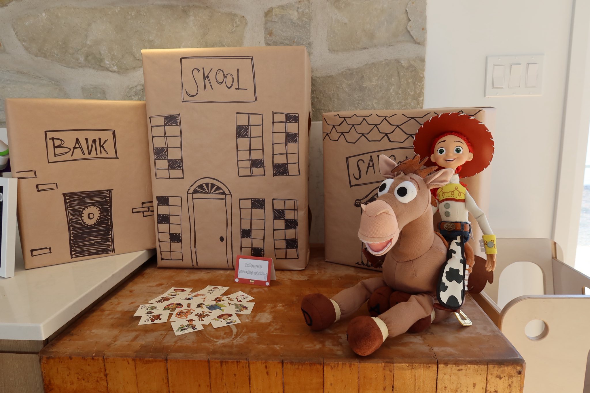 Jessie and Bullseye plush toys in front of cardboard box buildings labeled Skool, Bank, and Saloon.