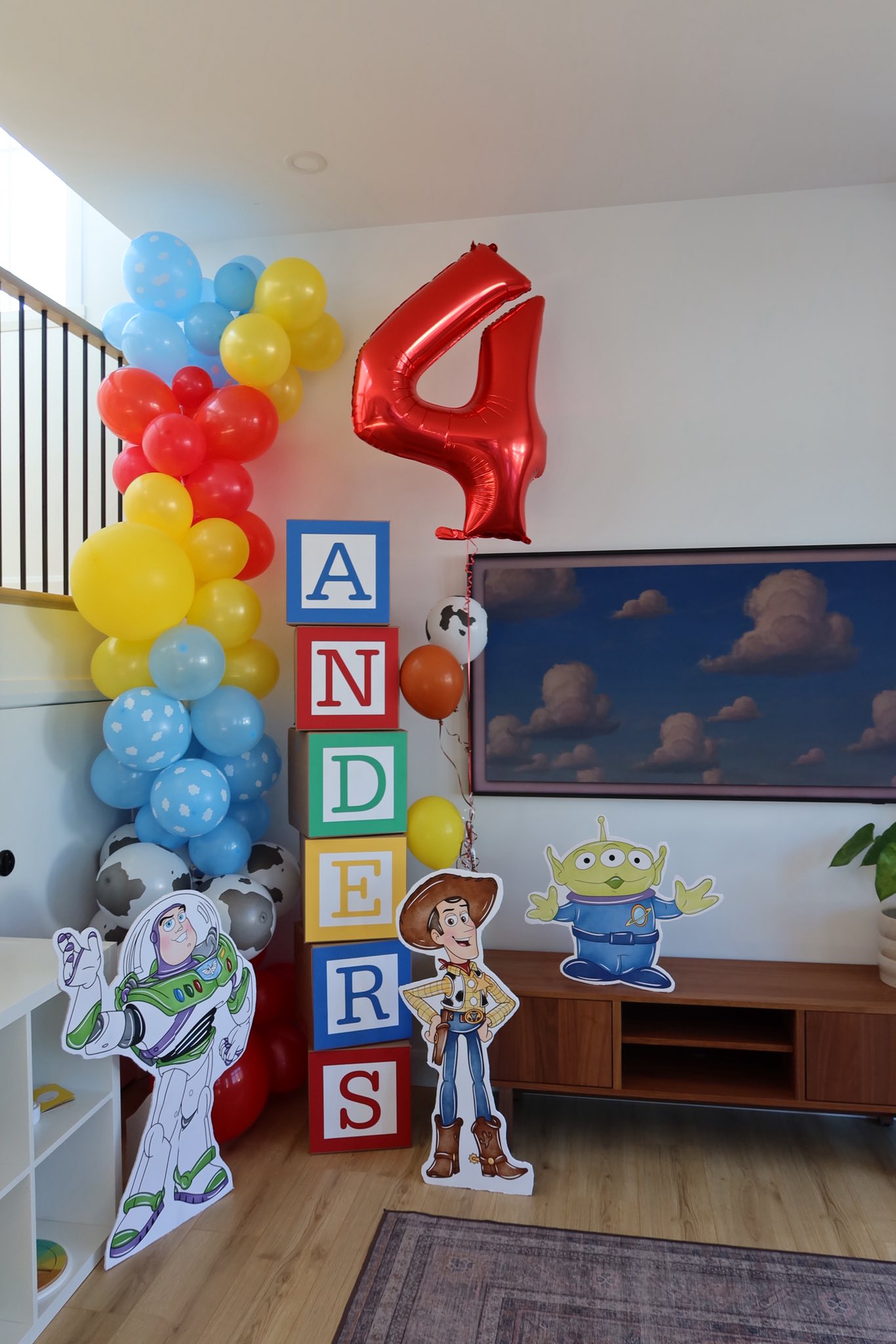Toy Story themed birthday party setup with Buzz Lightyear, Woody, and Alien cutouts, stacked letter blocks spelling ANDERS, and a red number four balloon.