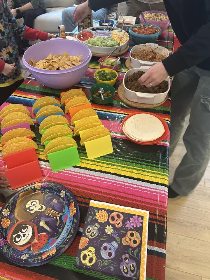 Taco bar spread on a striped Mexican tablecloth with taco shells, tortillas, seasoned meat, and various toppings.
