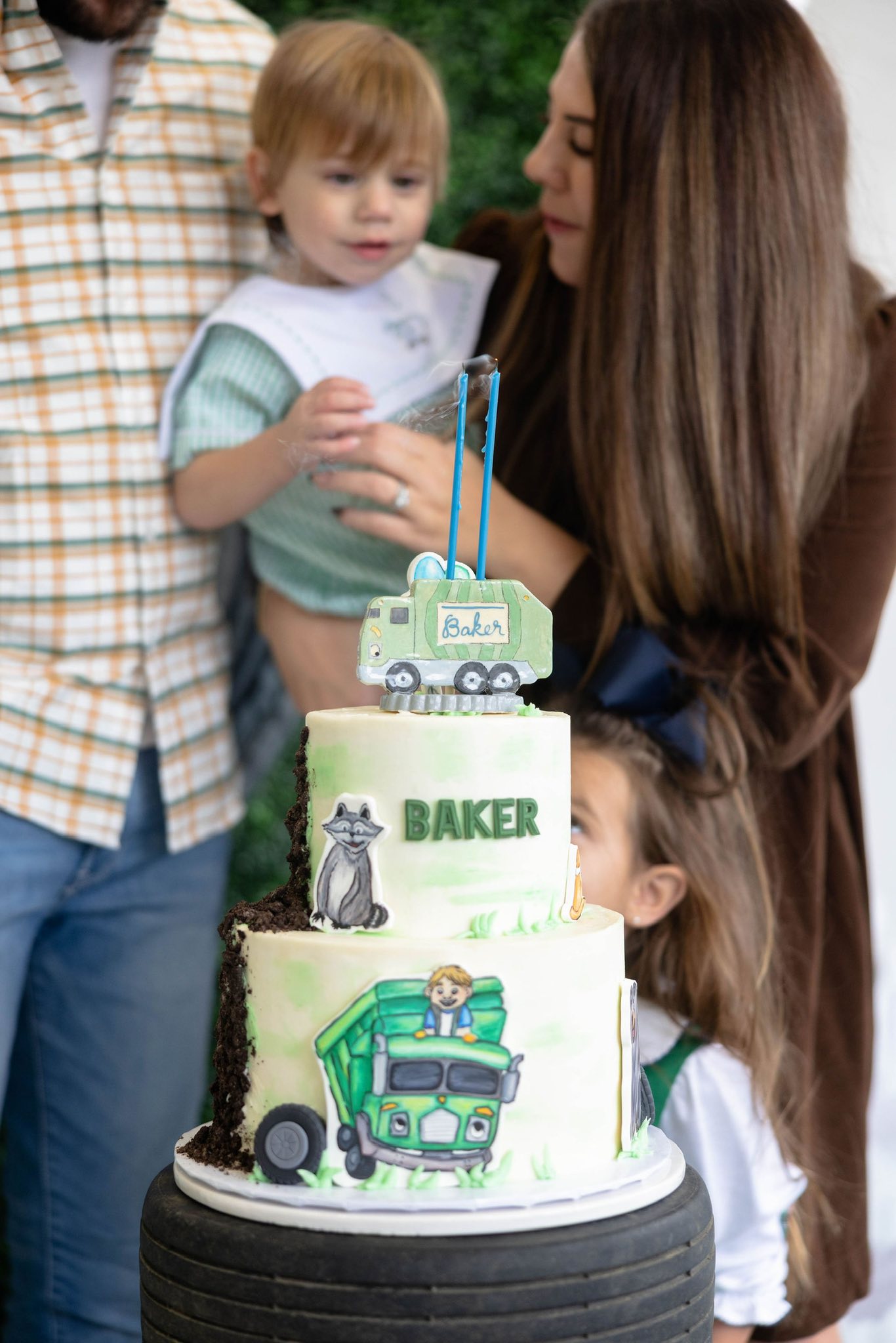 Two-tiered birthday cake with trash truck theme and Baker name label featuring a child being held by parents