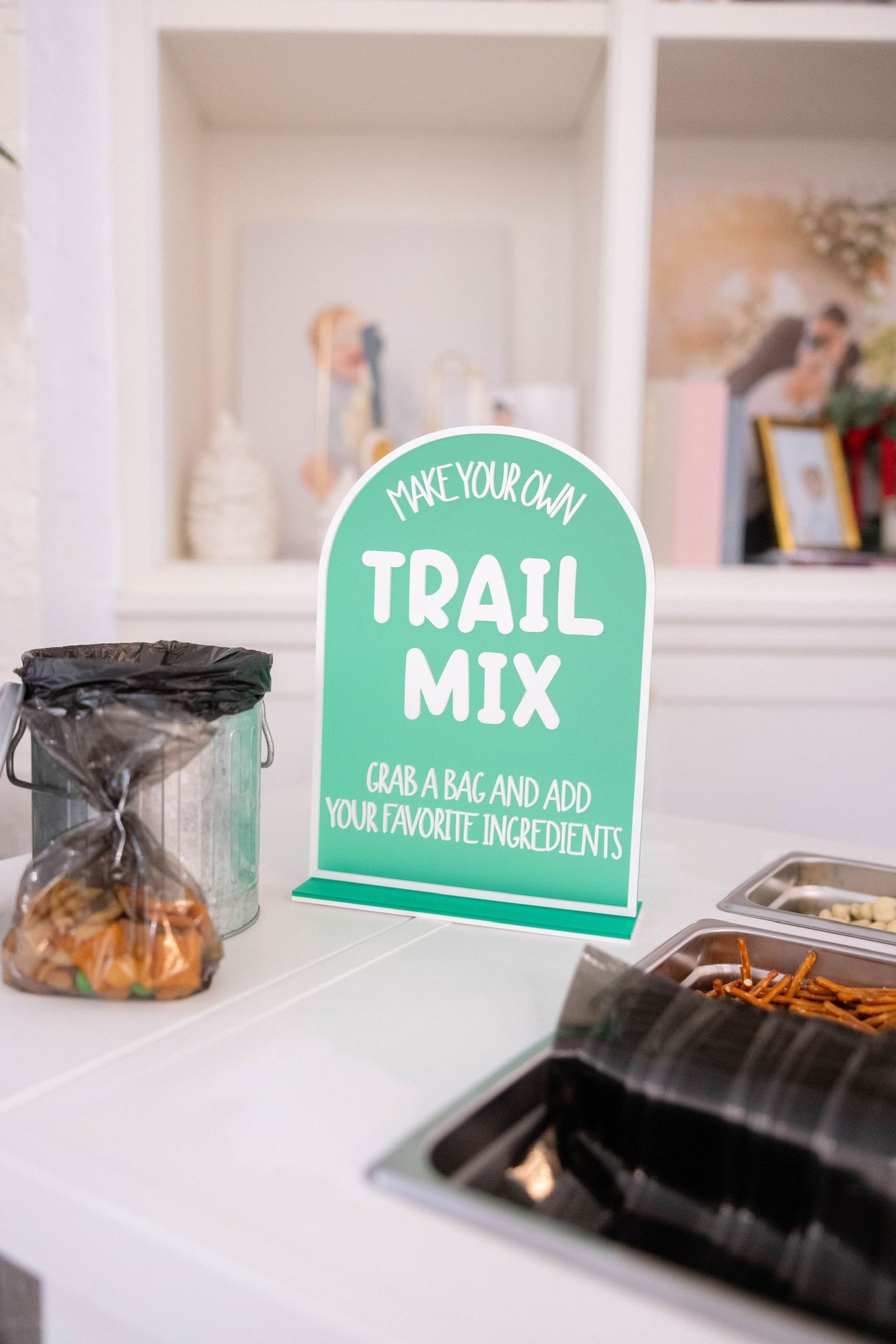 Table sign for a DIY trail mix bar with ingredients in trays and a container of bags.