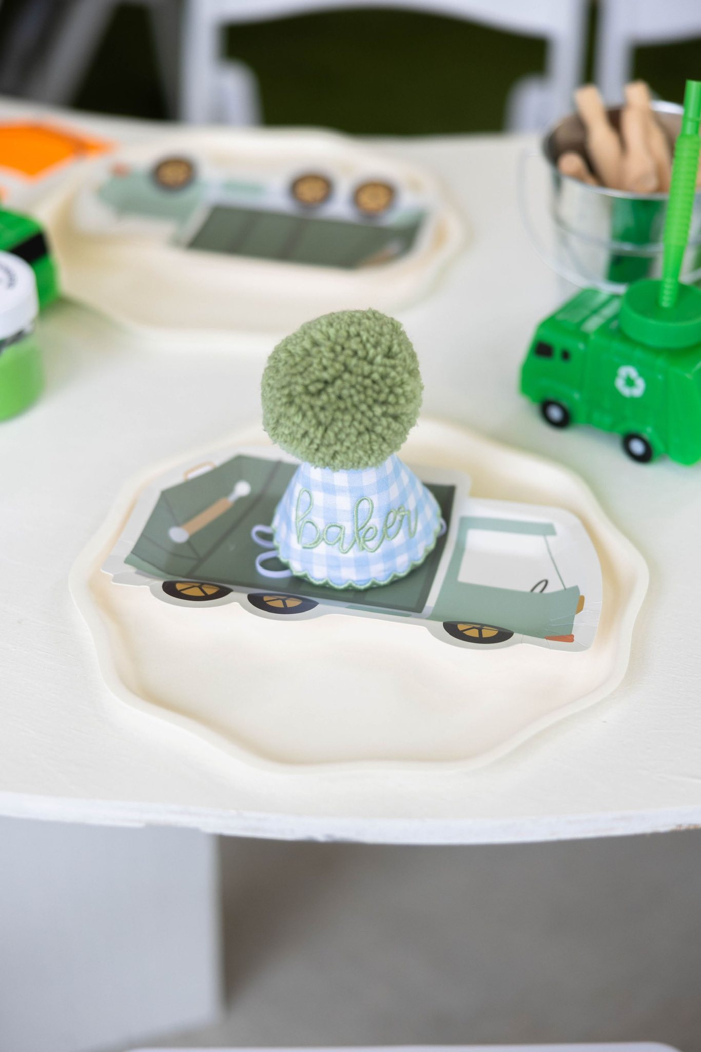 Personalized blue gingham party hat with a green pom-pom on top placed on a garbage truck shaped party plate.