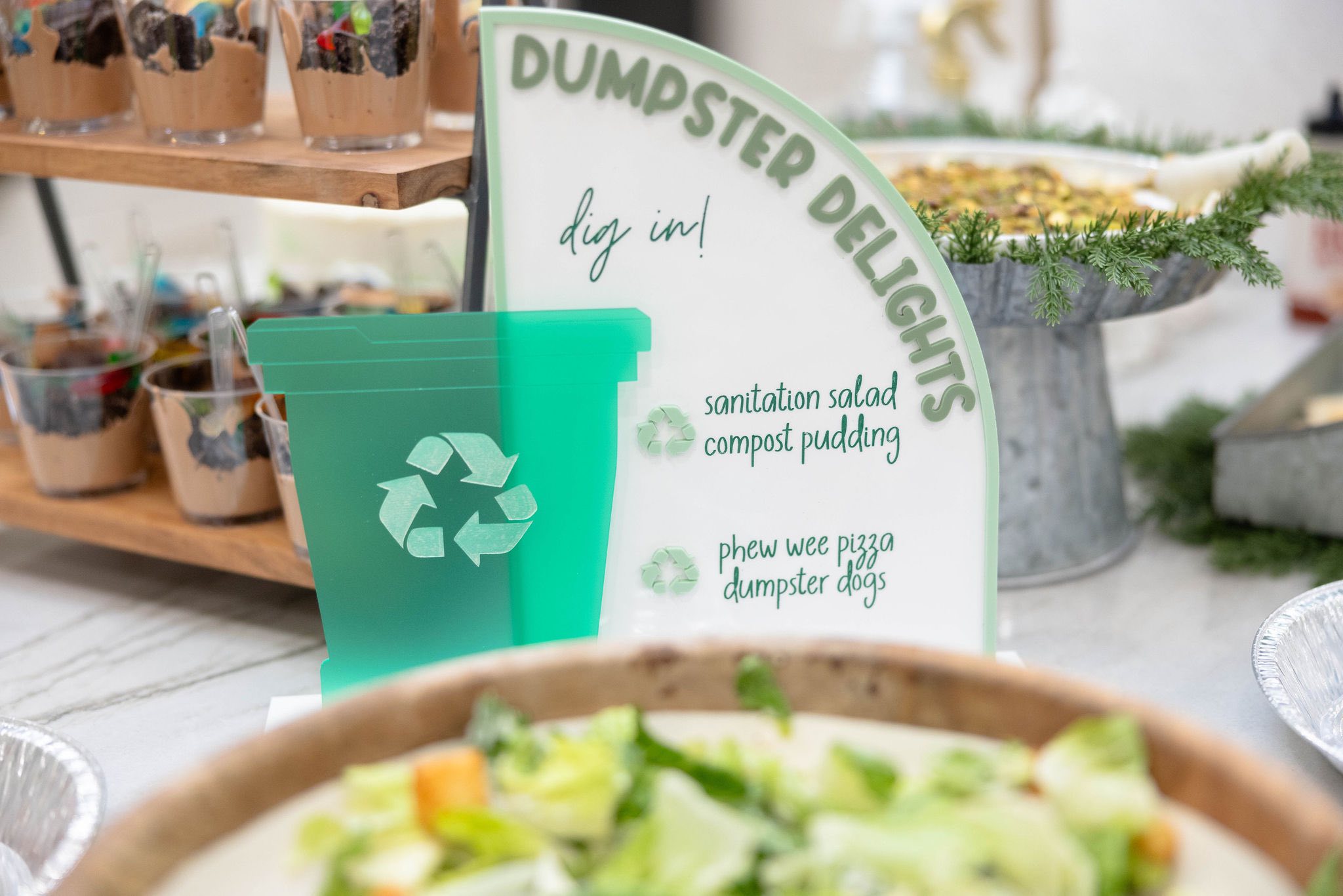 Table sign titled Dumpster Delights with a menu of themed party foods including compost pudding and dumpster dogs.