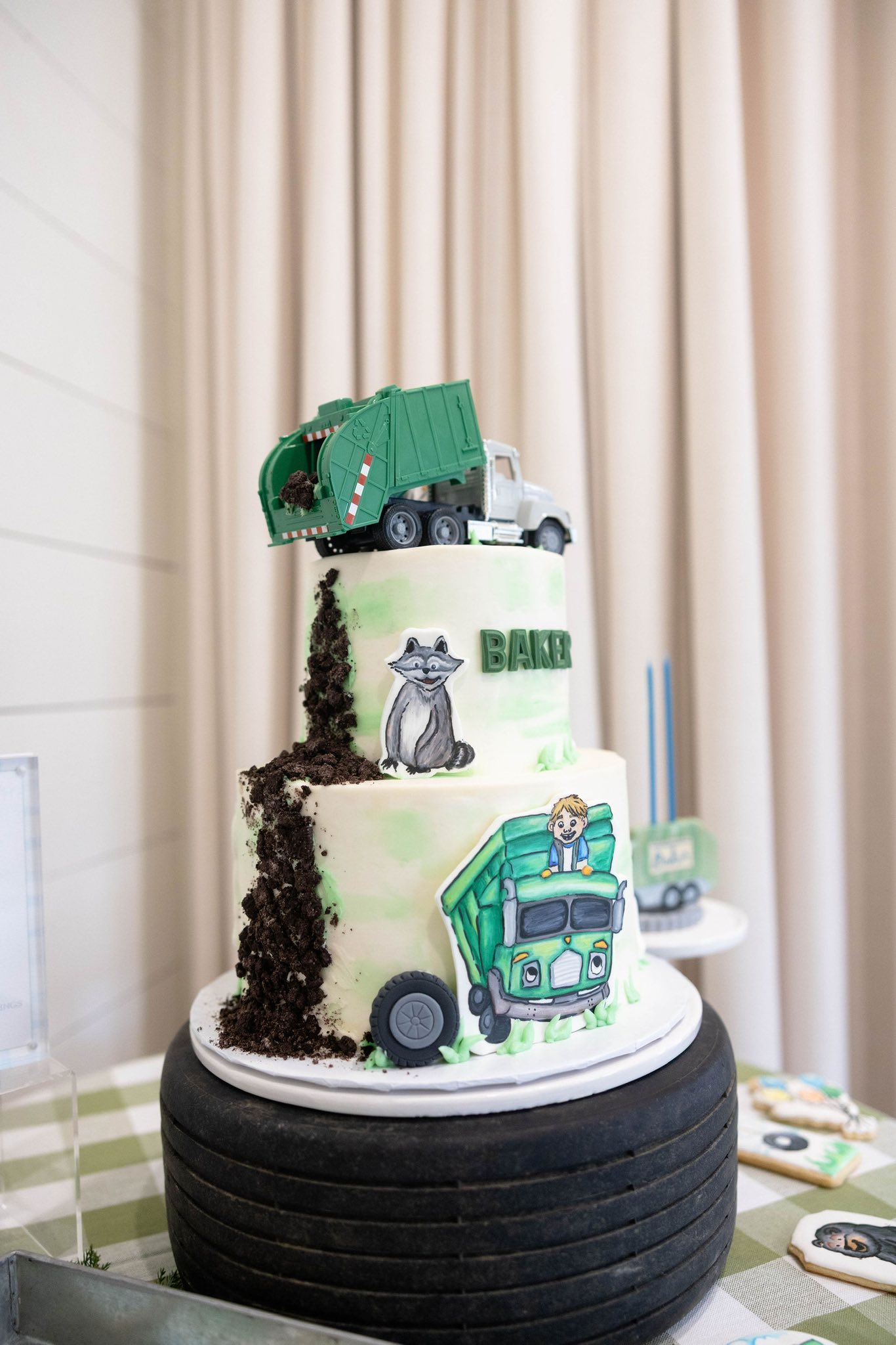 Two-tiered garbage truck themed birthday cake with a raccoon decoration and edible cookie crumbs