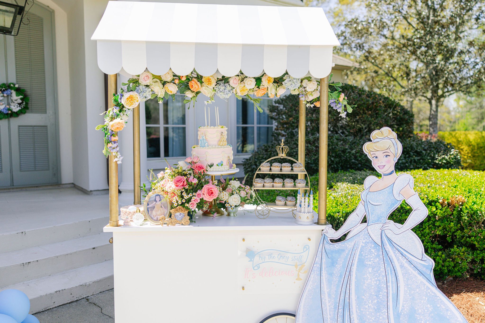 Cinderella themed party dessert cart with a tiered cake, floral arrangement, and cupcakes next to a Cinderella cutout