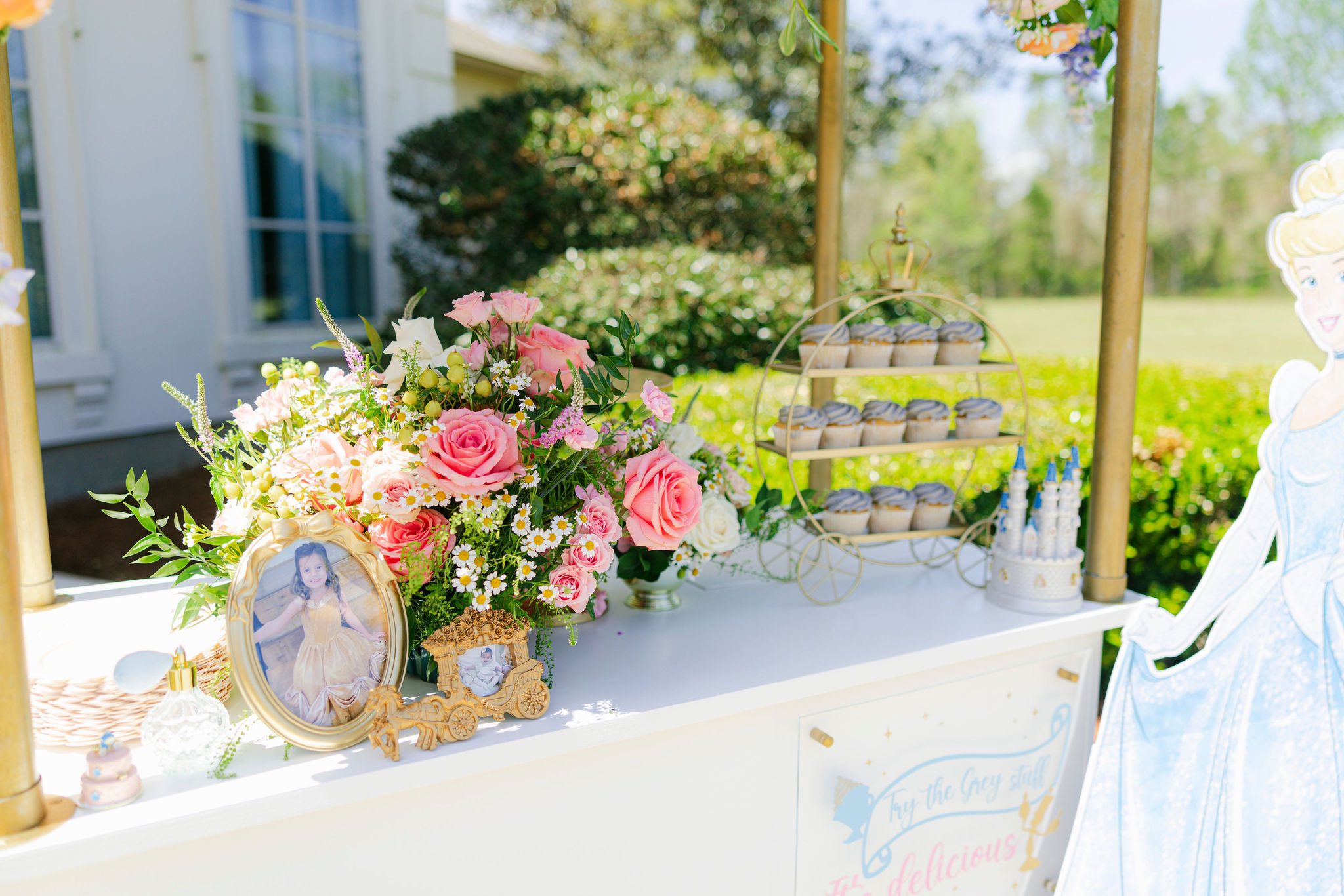 Cinderella-themed birthday party dessert cart with cupcakes, floral arrangement, and framed photos.