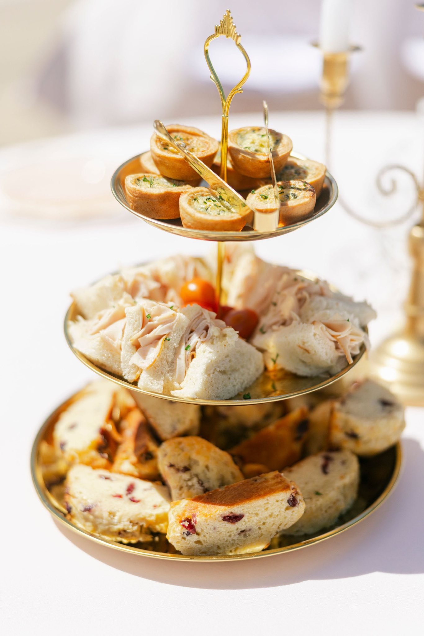 Three-tiered gold serving tray filled with tea sandwiches, savory mini quiches, and cranberry scones.