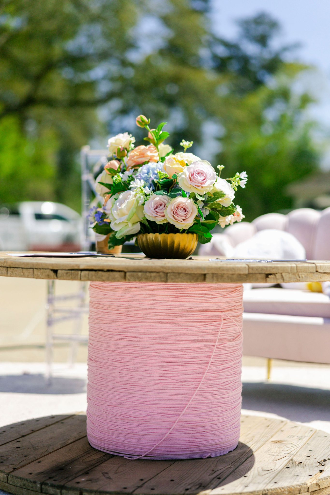 Table centerpiece of pastel roses and blue hydrangeas in a gold bowl on a pink yarn-wrapped spool table