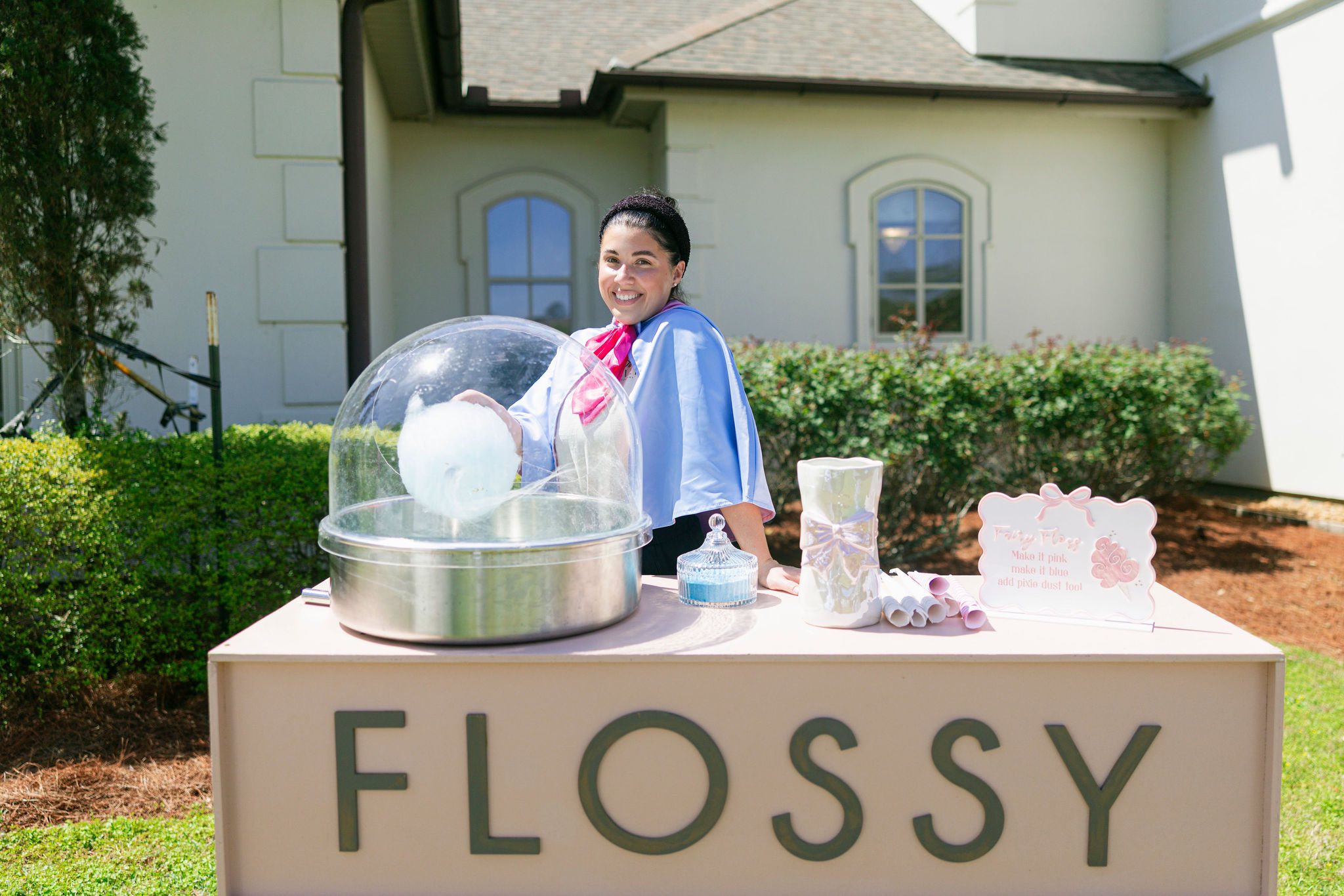 Woman standing behind a Flossy candy floss cart making blue cotton candy outdoors.