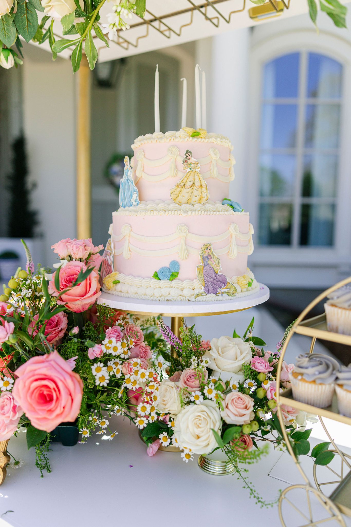 Two-tier pink princess birthday cake with Disney princess toppers surrounded by floral arrangements.