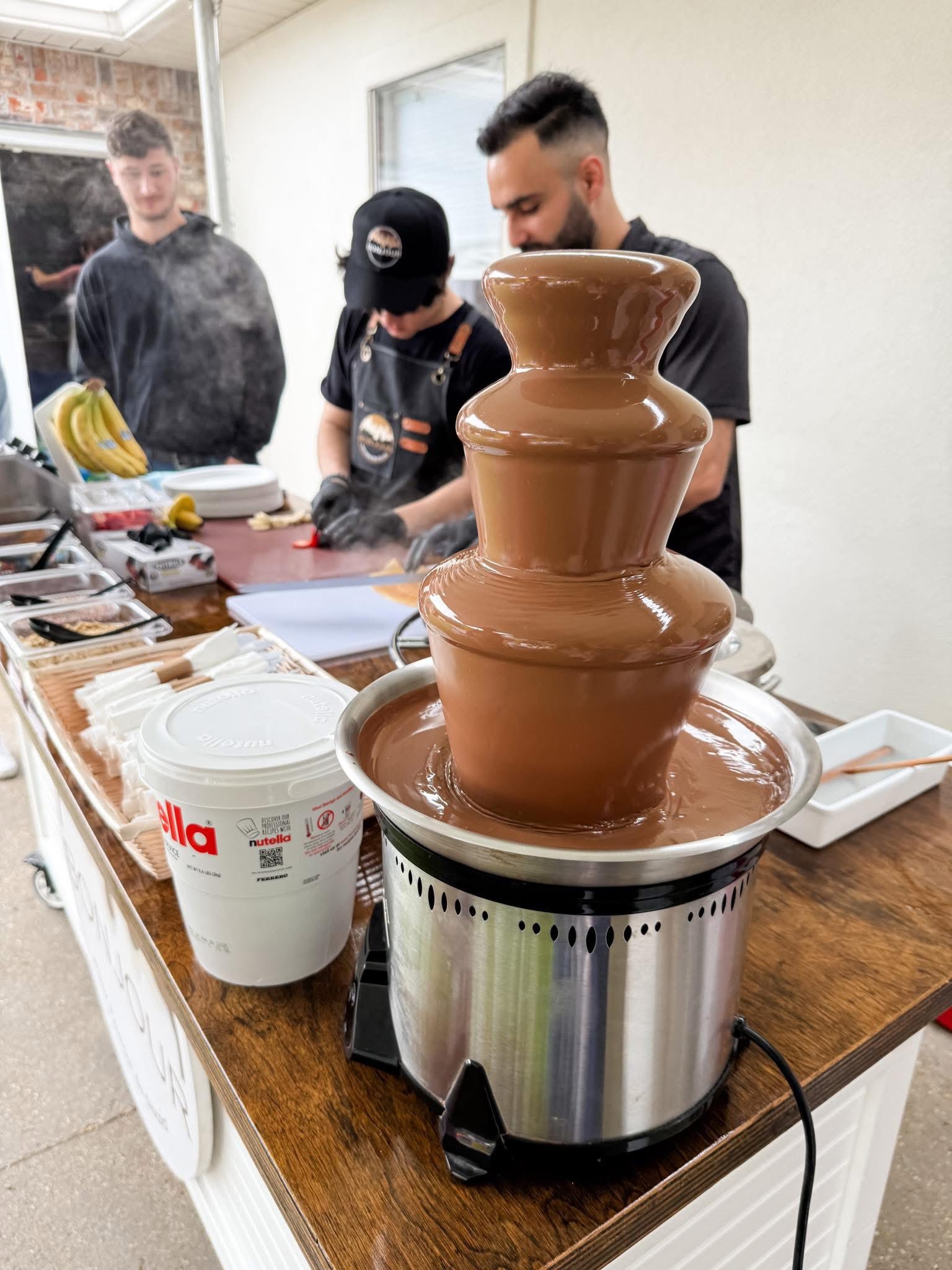 Tabletop chocolate fountain overflowing with melted Nutella next to a food preparation station.