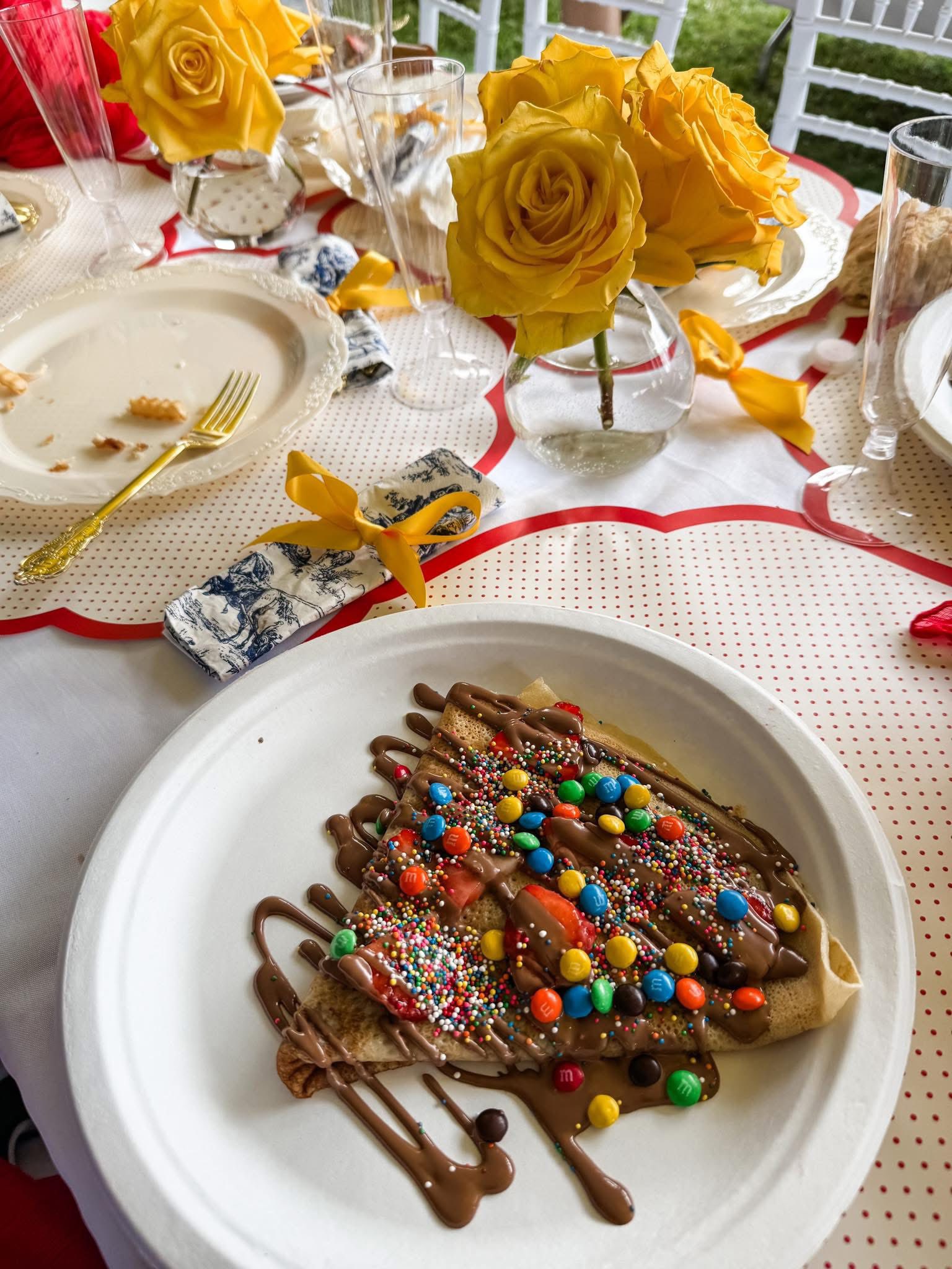 A crepe drizzled with chocolate sauce, topped with strawberry slices and colorful chocolate candies on a party table.
