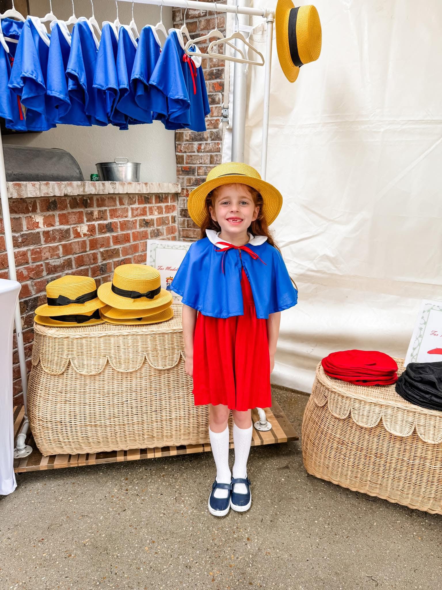 Young girl smiling wearing a Madeline costume including a red dress, blue cape, and yellow straw hat.