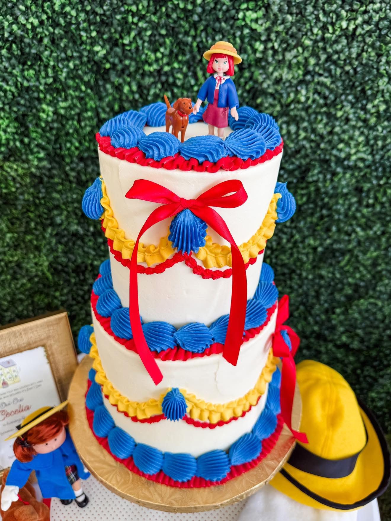 Two-tier white cake decorated with red, yellow, and blue buttercream piping, red ribbon, and Madeline figure topper.