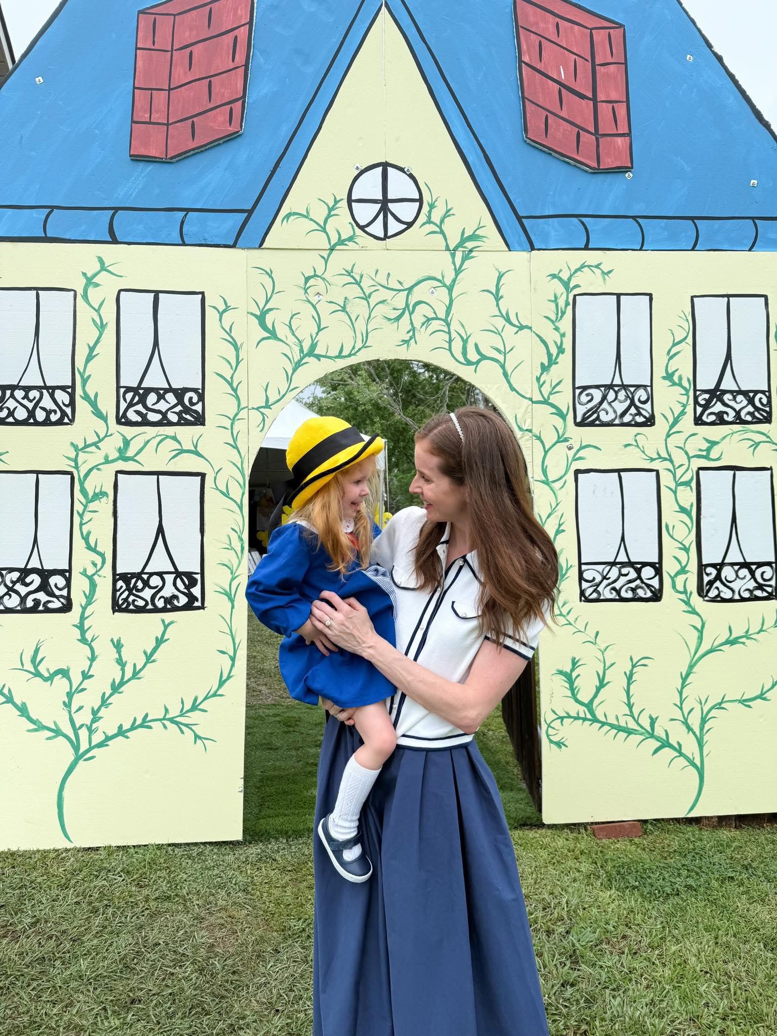 Mother holding her young daughter dressed in a Madeline inspired blue costume and yellow hat in front of a hand-painted house cutout.
