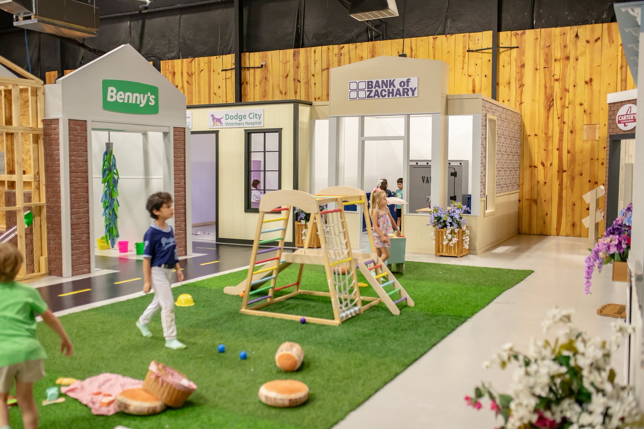 Children playing in an indoor miniature town featuring a bank, vet, and car wash with a wooden climbing frame.