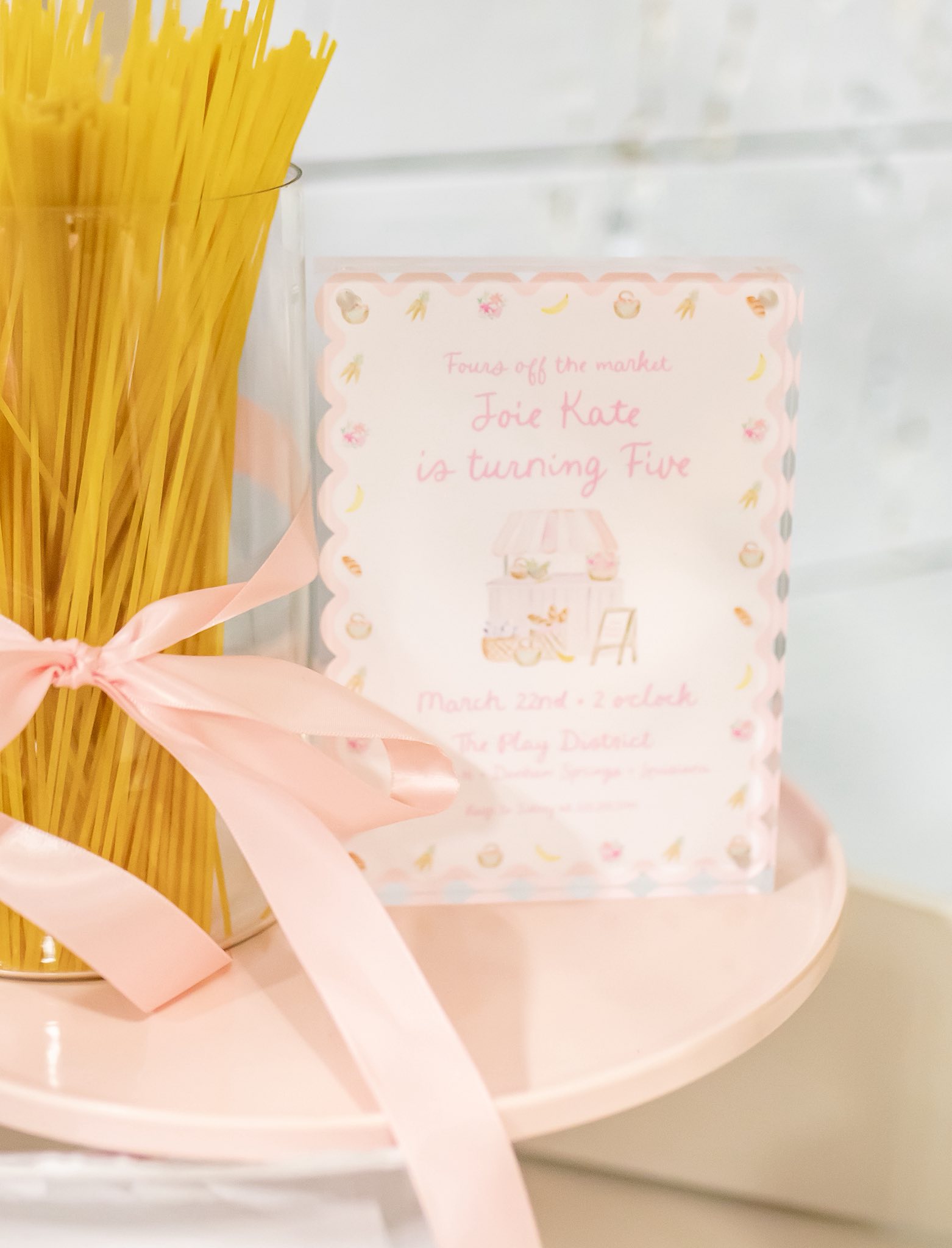Farmers market themed fifth birthday party invitation displayed on a pink tray with spaghetti and a pink ribbon.
