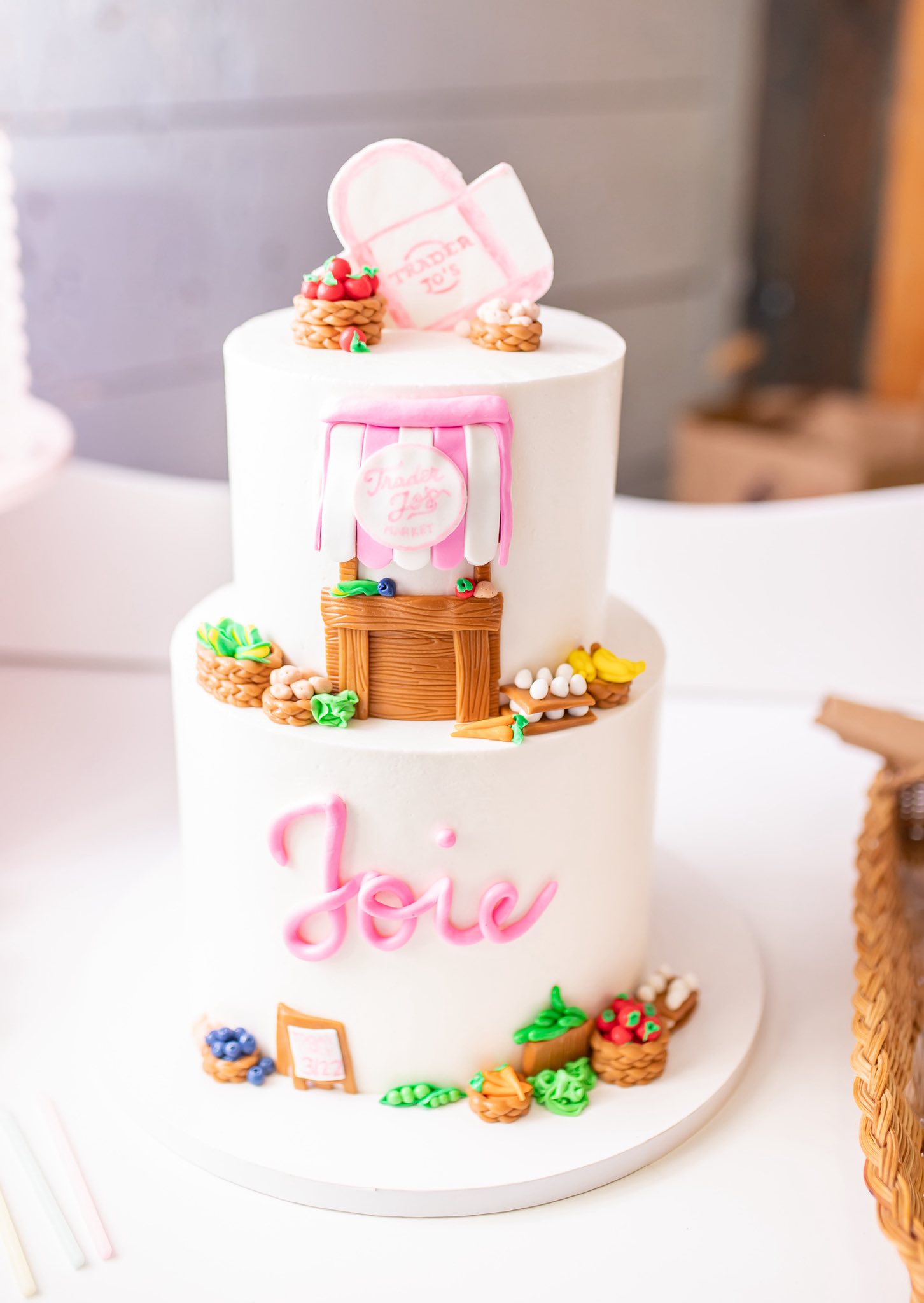 Two-tier white cake decorated with a Trader Joe's market theme and the name Joie in pink script.