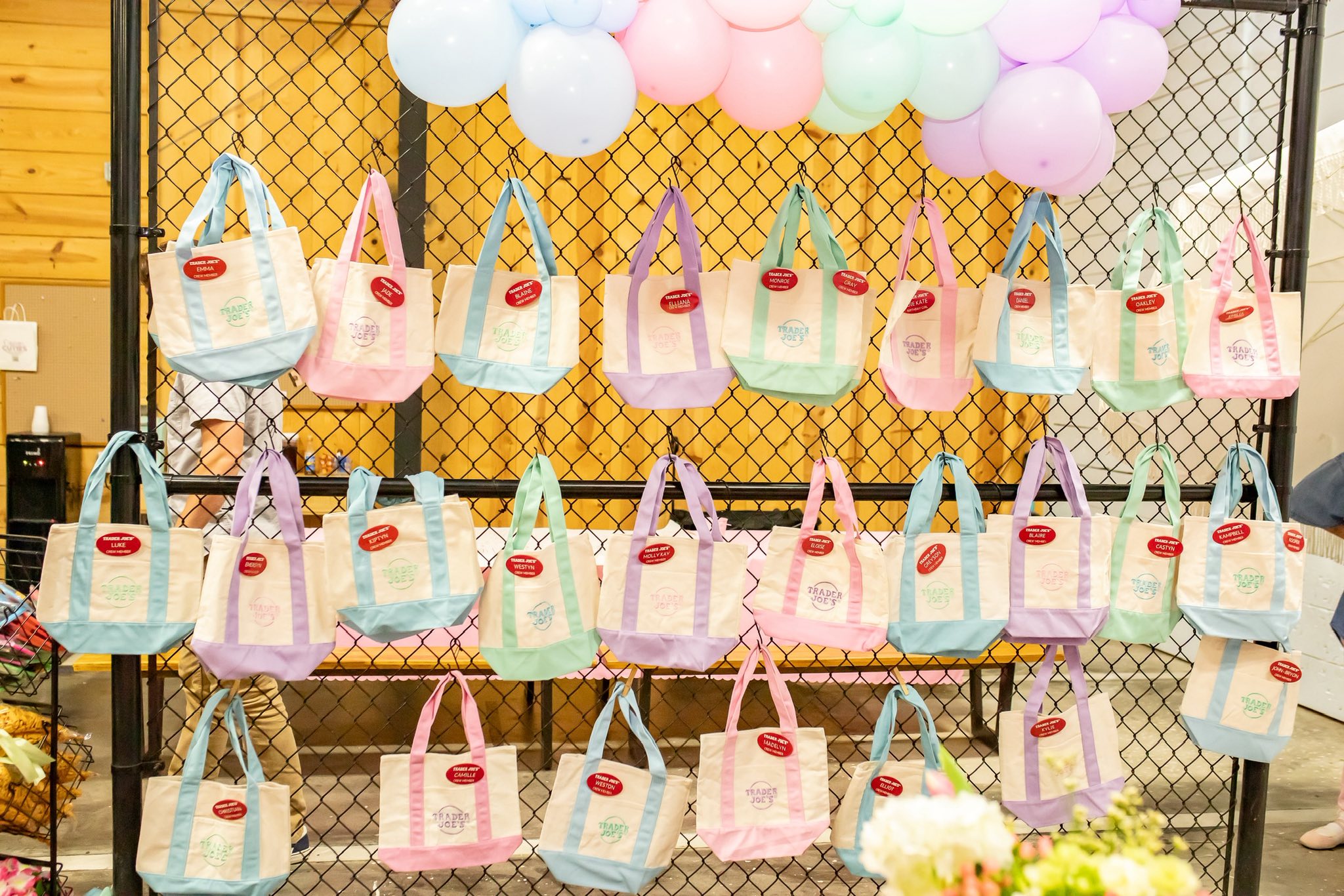Display of colorful canvas tote bags featuring the Trader Joe's logo hanging on a wire fence.