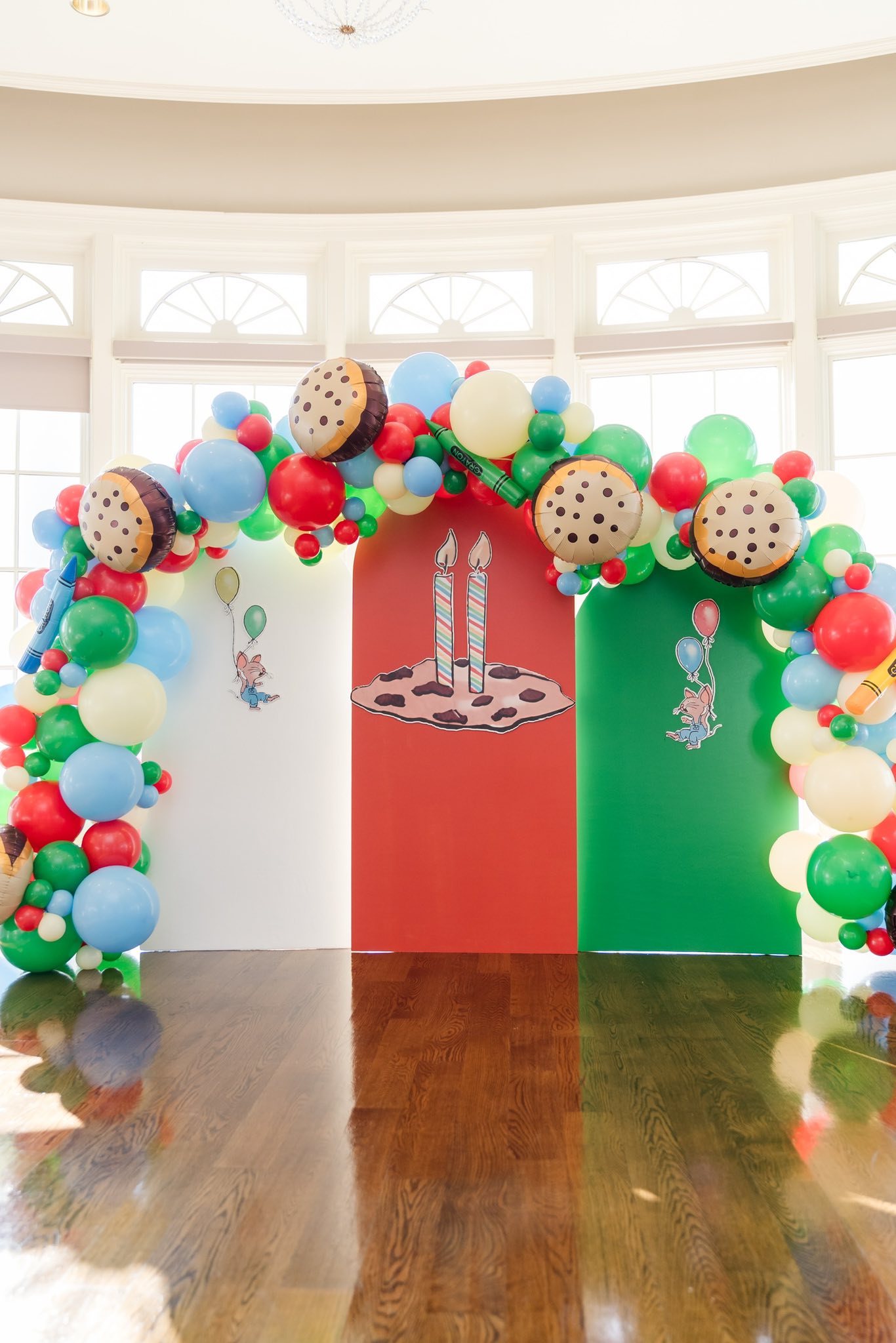 Colorful balloon arch with cookie-themed party decorations and mouse illustrations against arched wall panels.