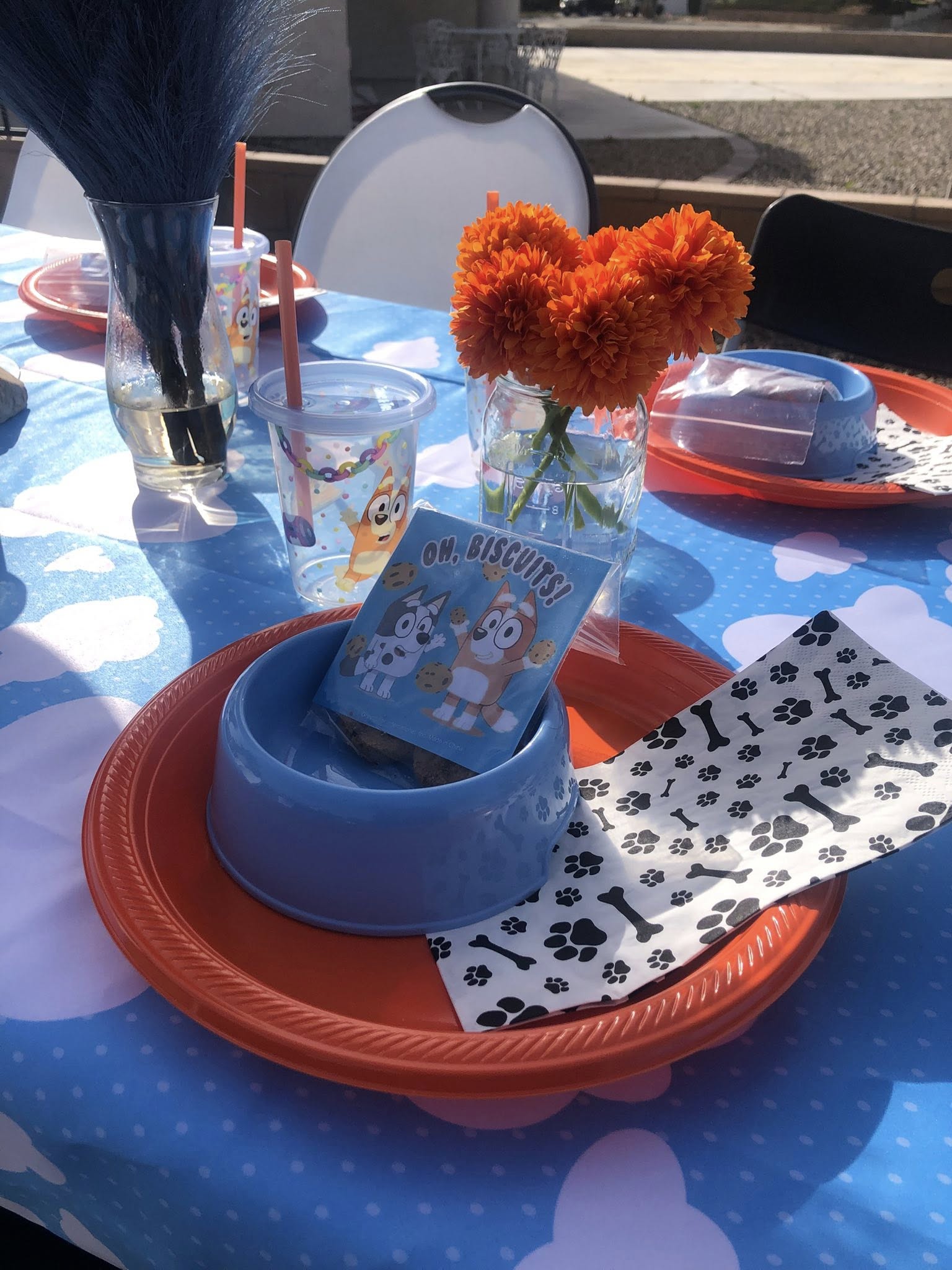 Bluey-themed party place setting with a light blue bowl, paw print napkin, and biscuit bag on an orange plate.