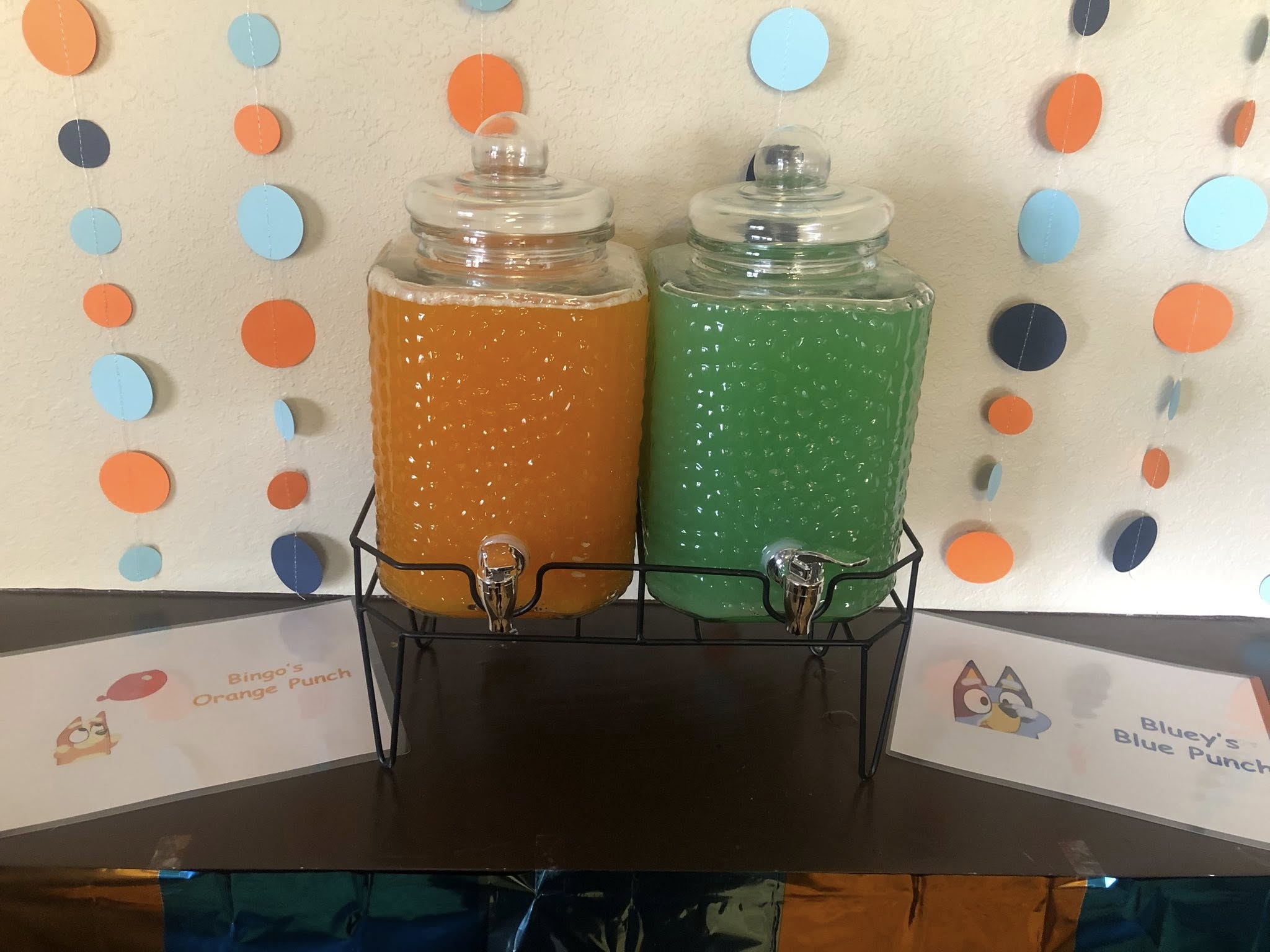 Two glass beverage dispensers labeled as Bingo's orange punch and Bluey's blue punch at a party.
