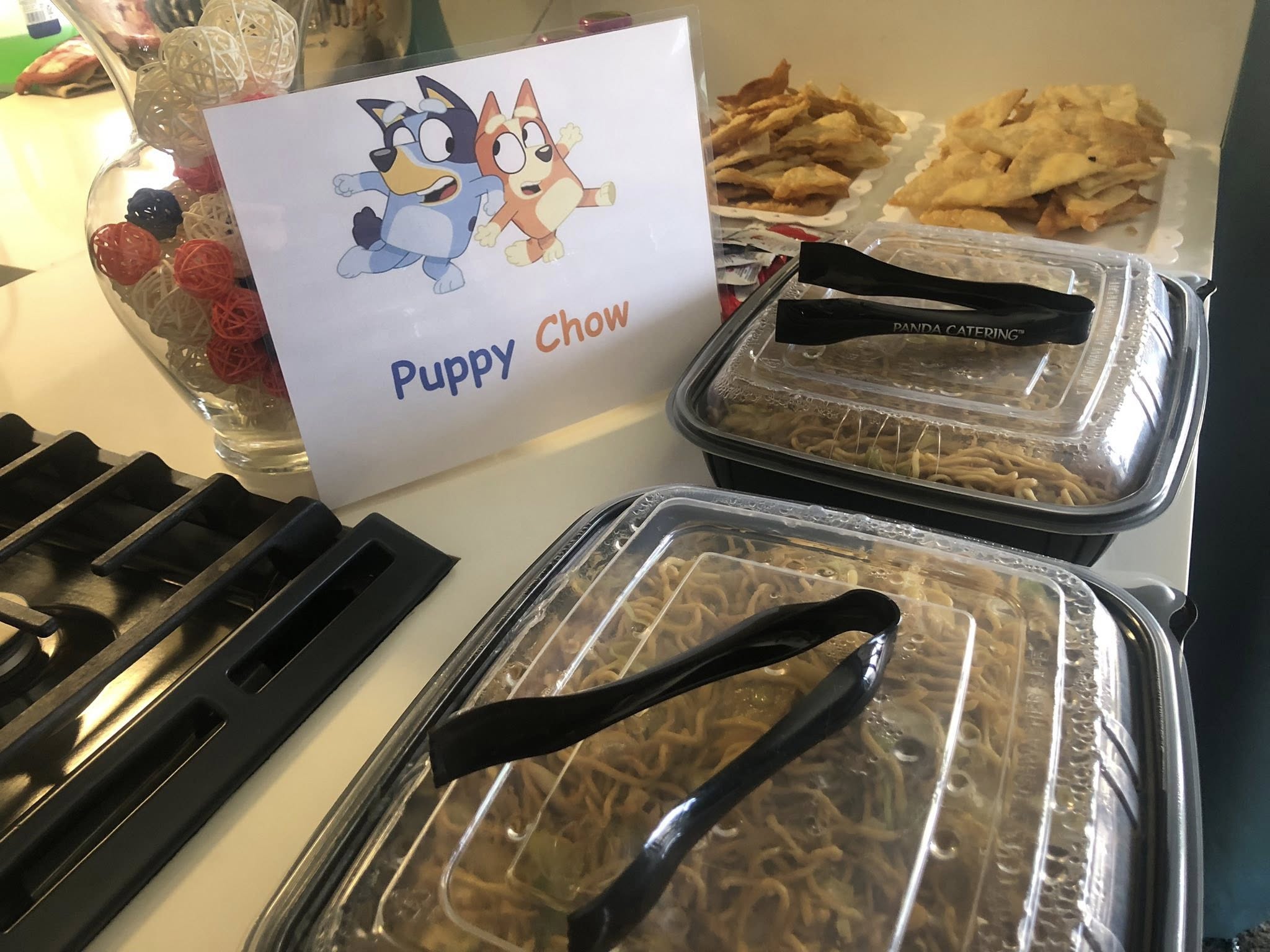Catering trays of chow mein labeled with a Bluey-themed Puppy Chow sign at a kid's party.
