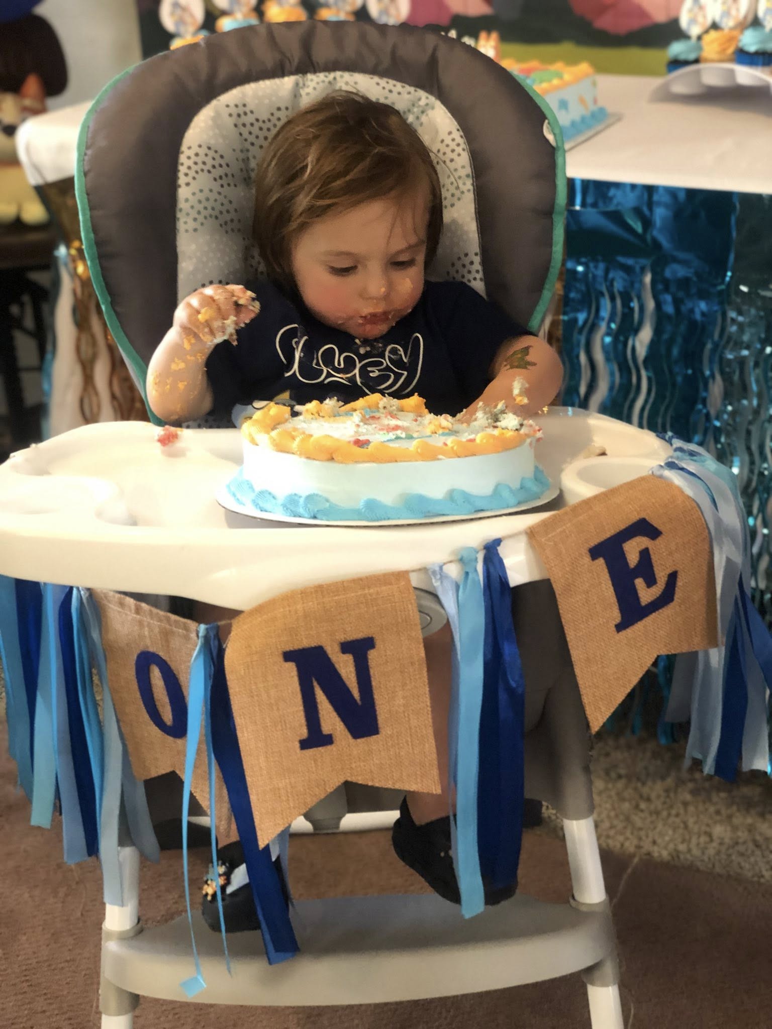 One-year-old child wearing a first birthday shirt messy with frosting while sitting in a high chair with a blue cake