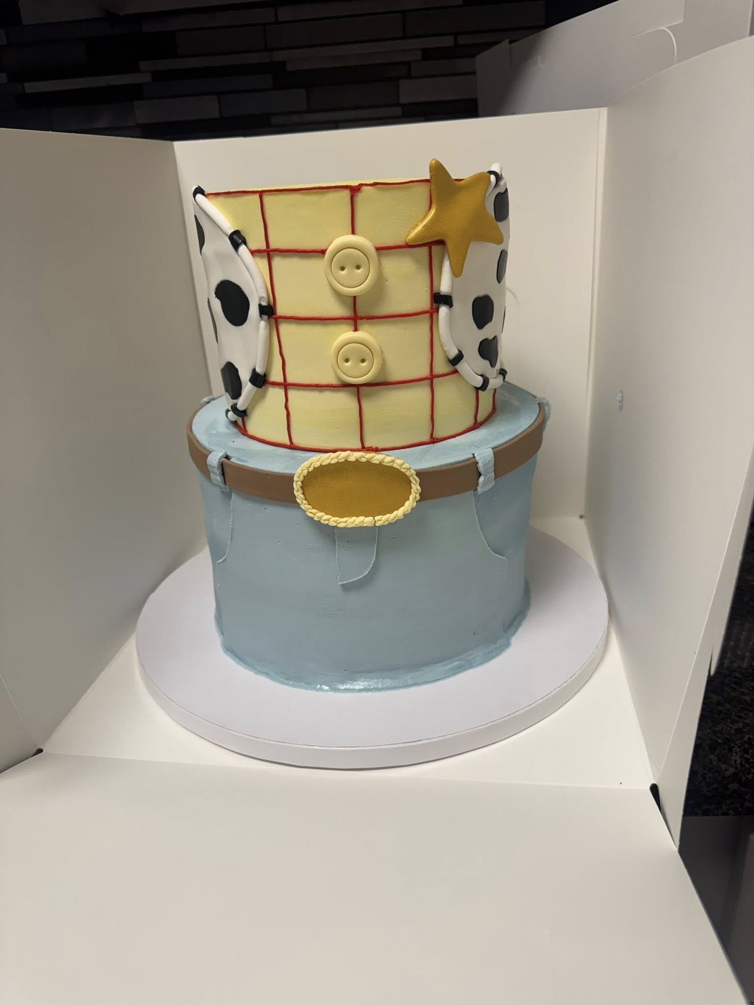 Two-tiered Woody from Toy Story themed birthday cake with yellow plaid bodice, cow-print vest, and blue jeans base